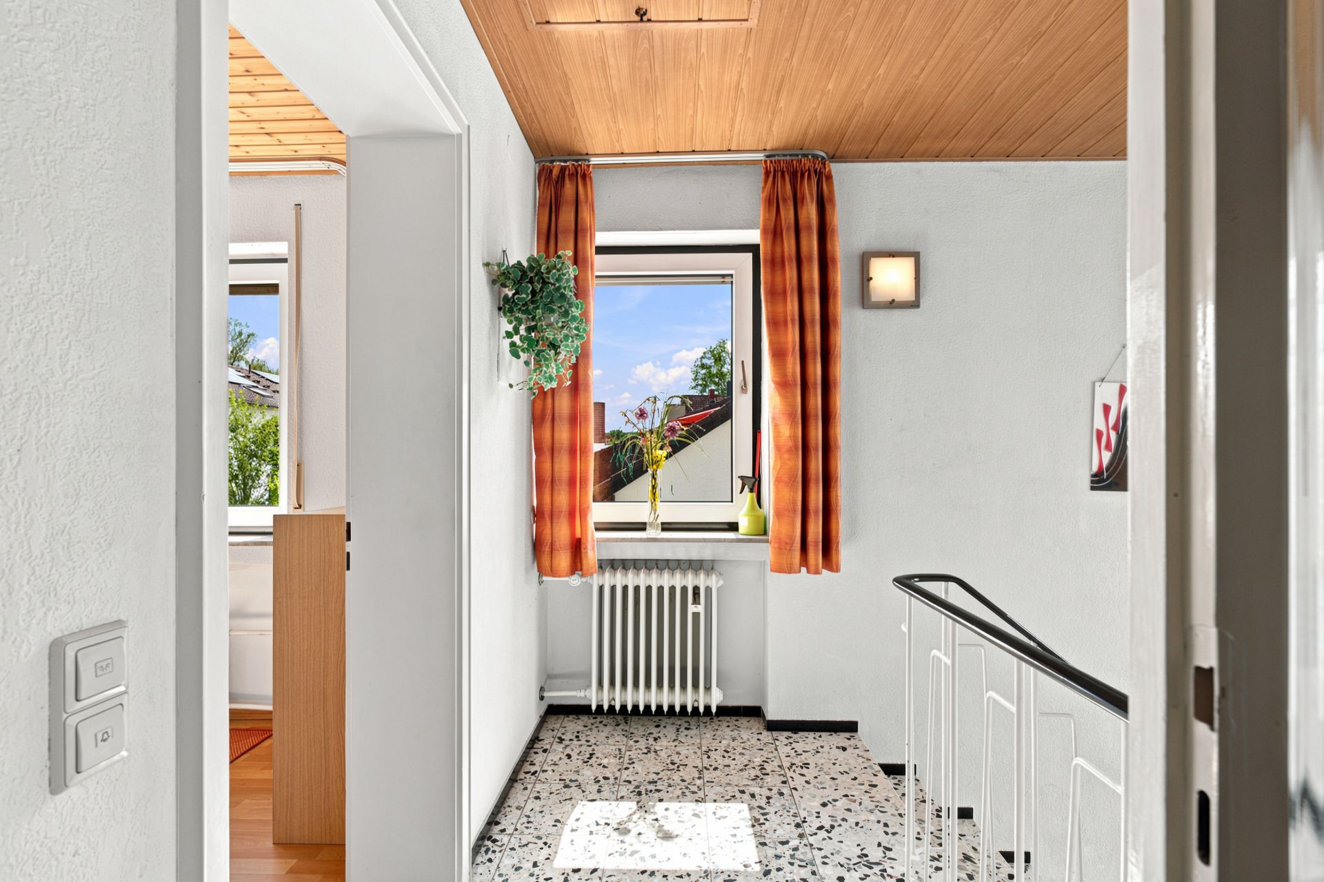 Heller Flur mit Fenster, orange Vorhängen, Treppengeländer und weißem Heizkörper.