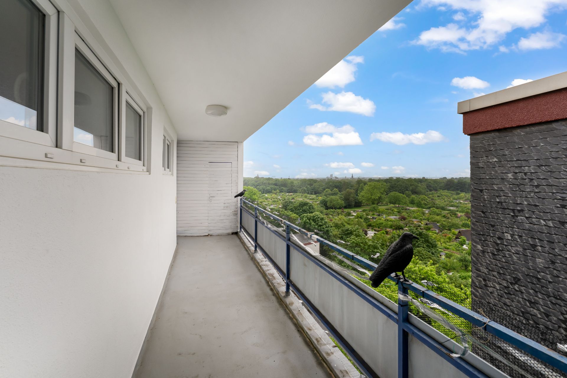Langer Balkon mit Geländer, Blick ins Grüne, daneben ein Gebäude, Himmel mit Wolken.