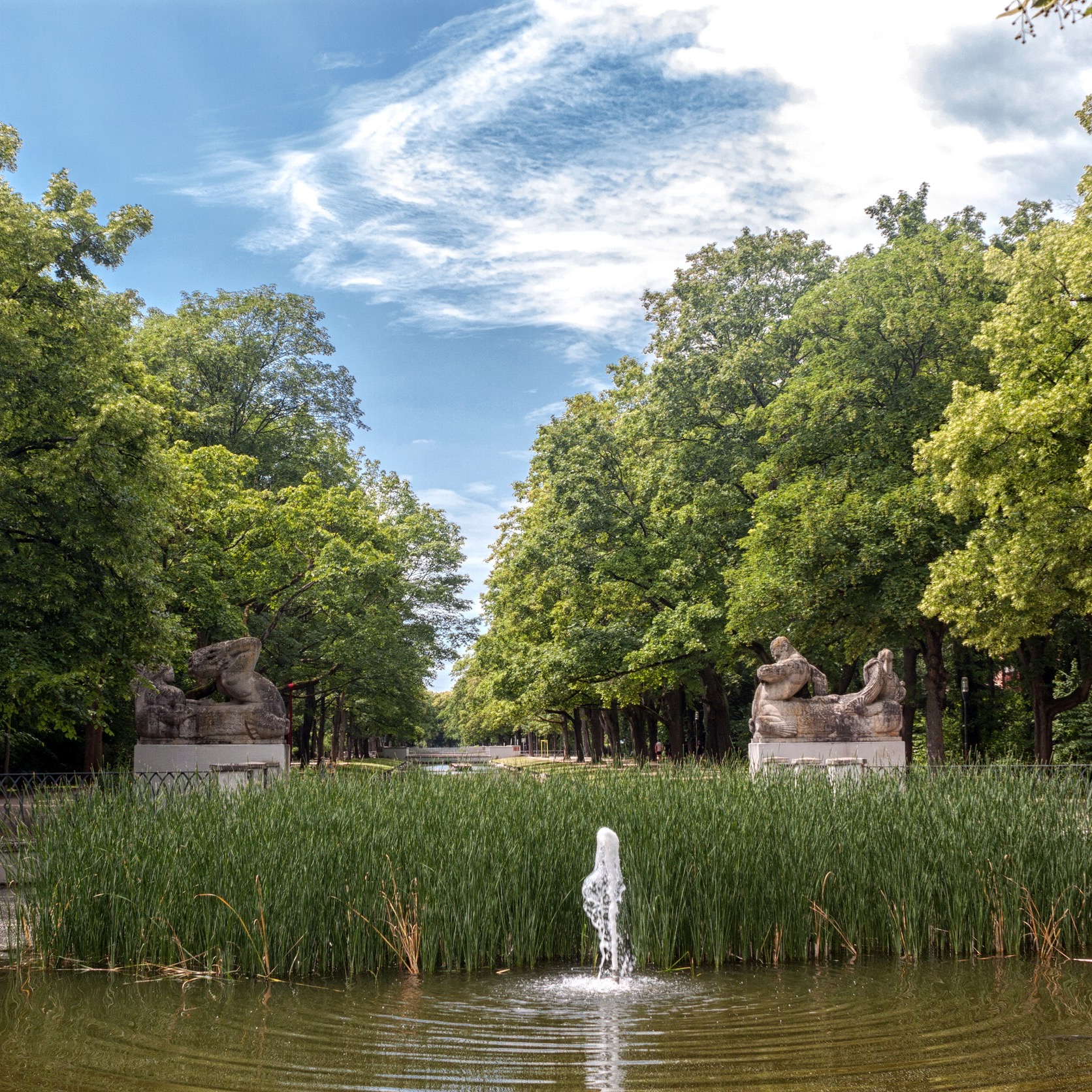Springbrunnen im Teich, umgeben von hohen Bäumen und zwei Steinskulpturen am Rand.