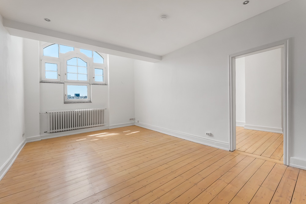 Helles, leeres Zimmer mit Holzboden, großem Fenster und Tür zu weiterem Raum.
