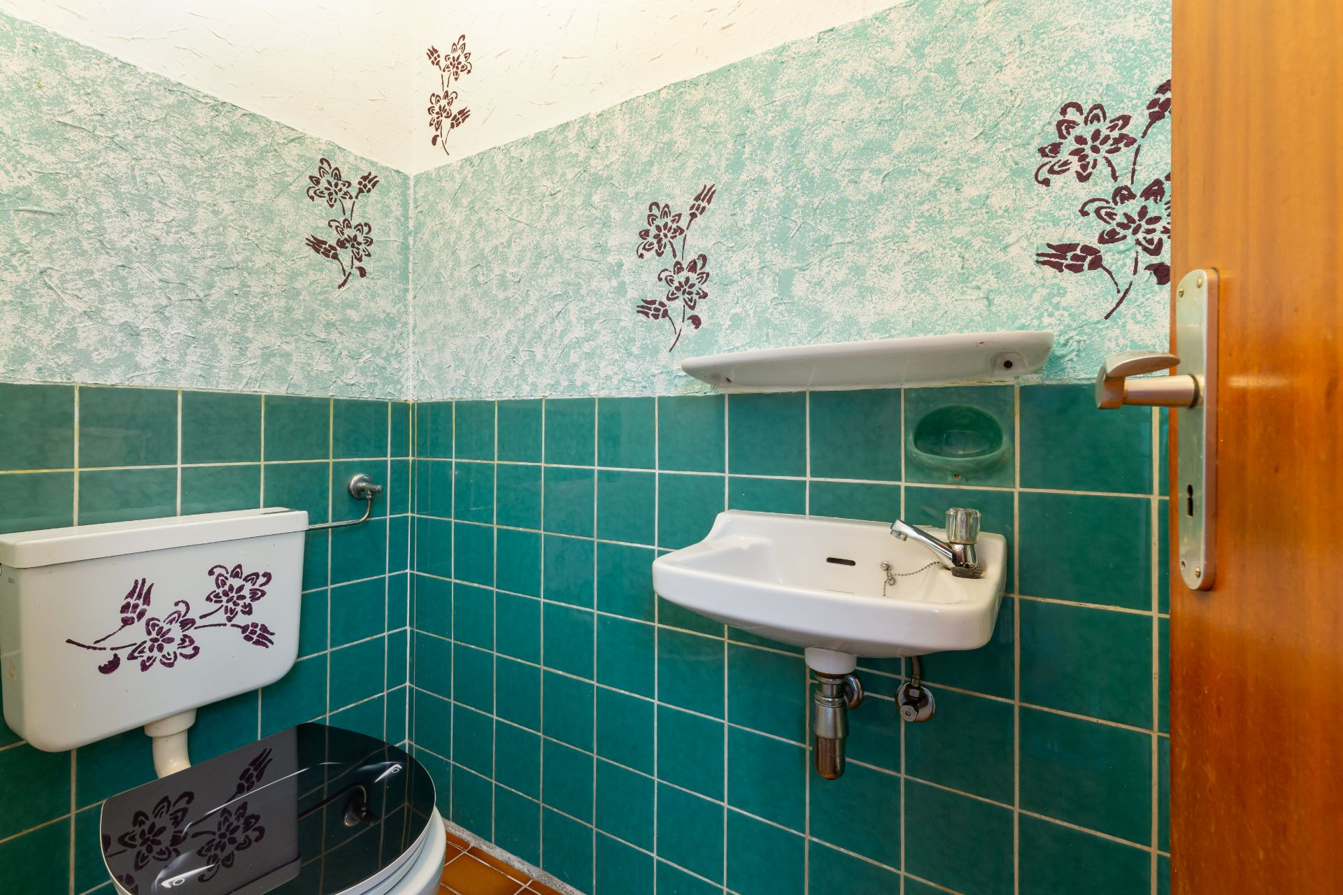 Kleines Badezimmer mit grün gefliesten Wänden, Toilette, Waschbecken und floralem Wanddekor.
