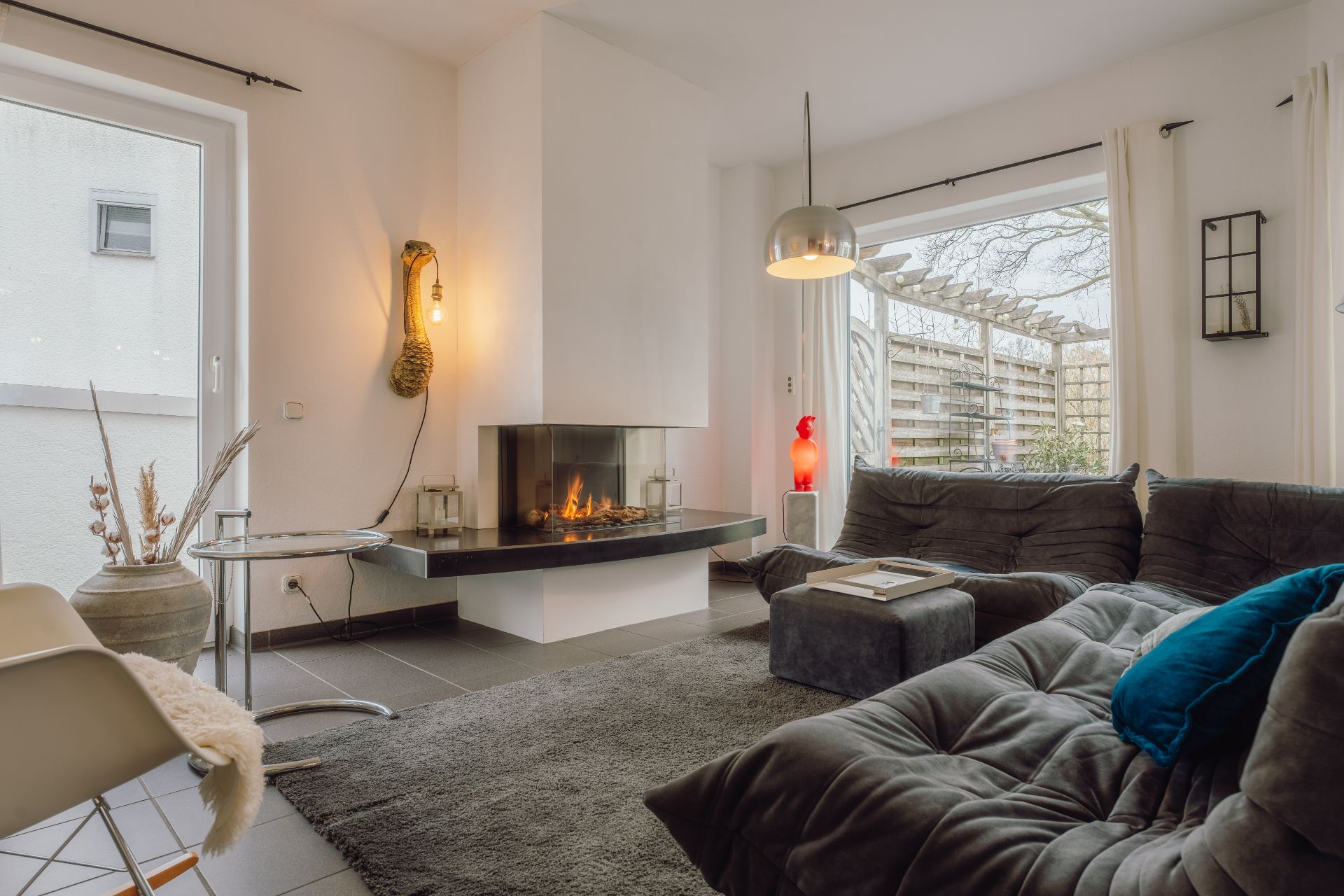 Modernes Wohnzimmer mit Kamin, grauem Sofa, Teppich und großen Fenstern zum Garten.
