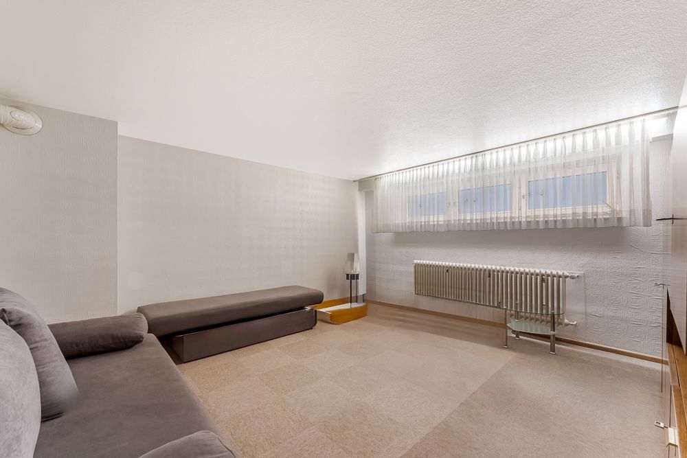 Helles Zimmer mit zwei Sofas, Heizung, Stehlampe und breitem Fenster mit Vorhang.