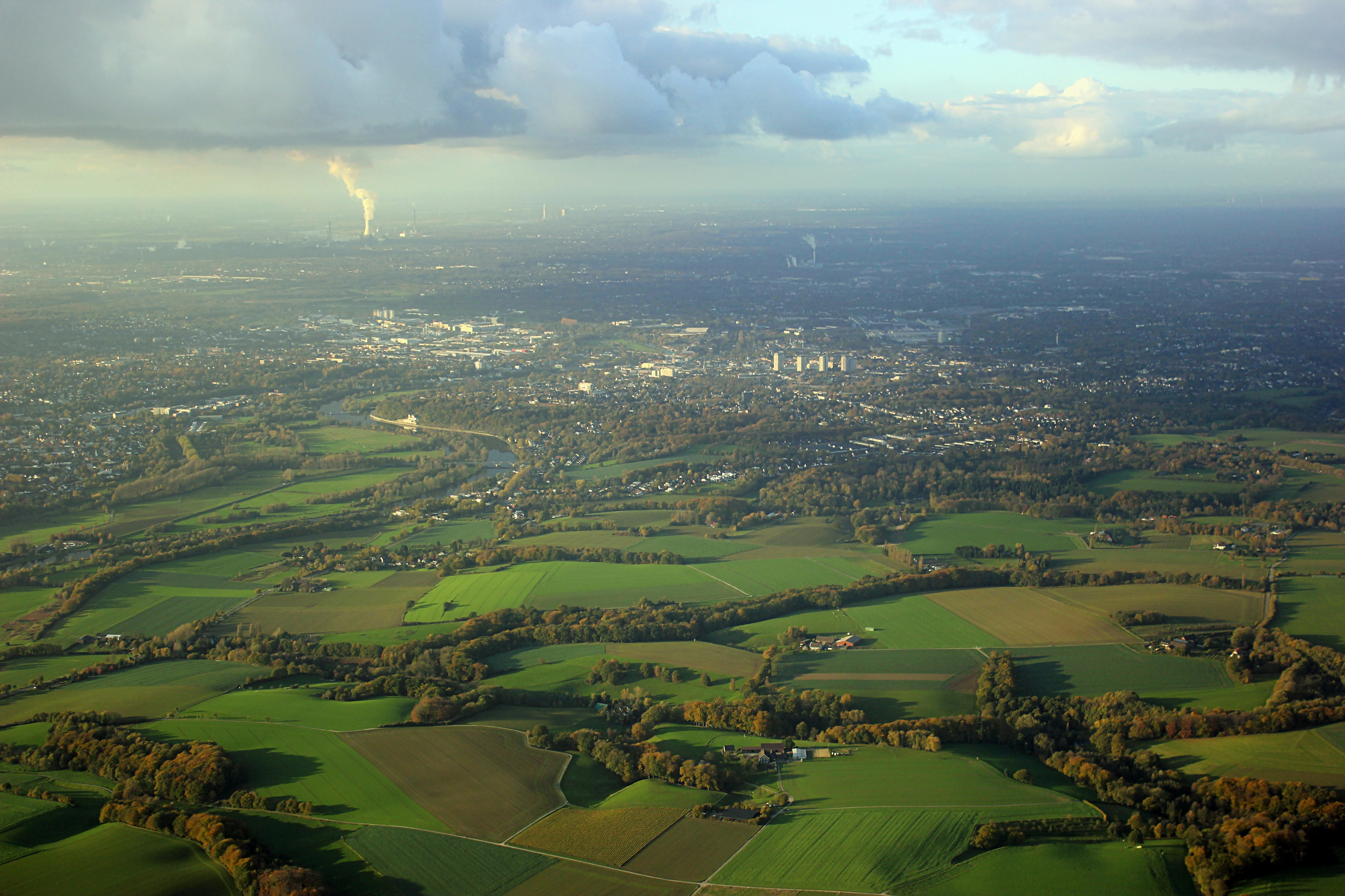 Luftaufnahme von grünen Feldern und einer Stadt im Hintergrund bei bewölktem Himmel.