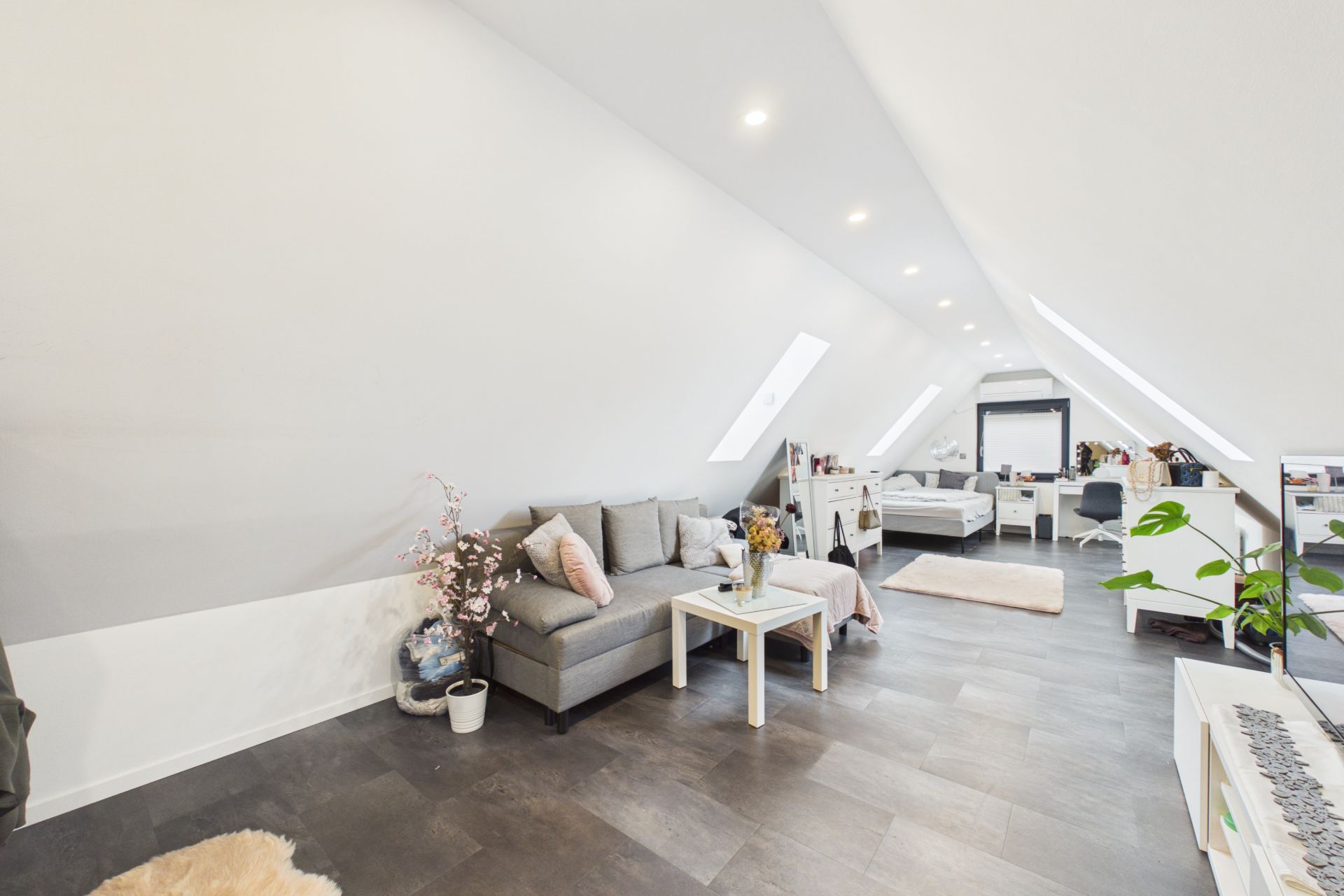 Heller Dachgeschossraum mit Sofa, Tisch, Bett und Schreibtisch unter schrägen Decken und Dachfenstern.