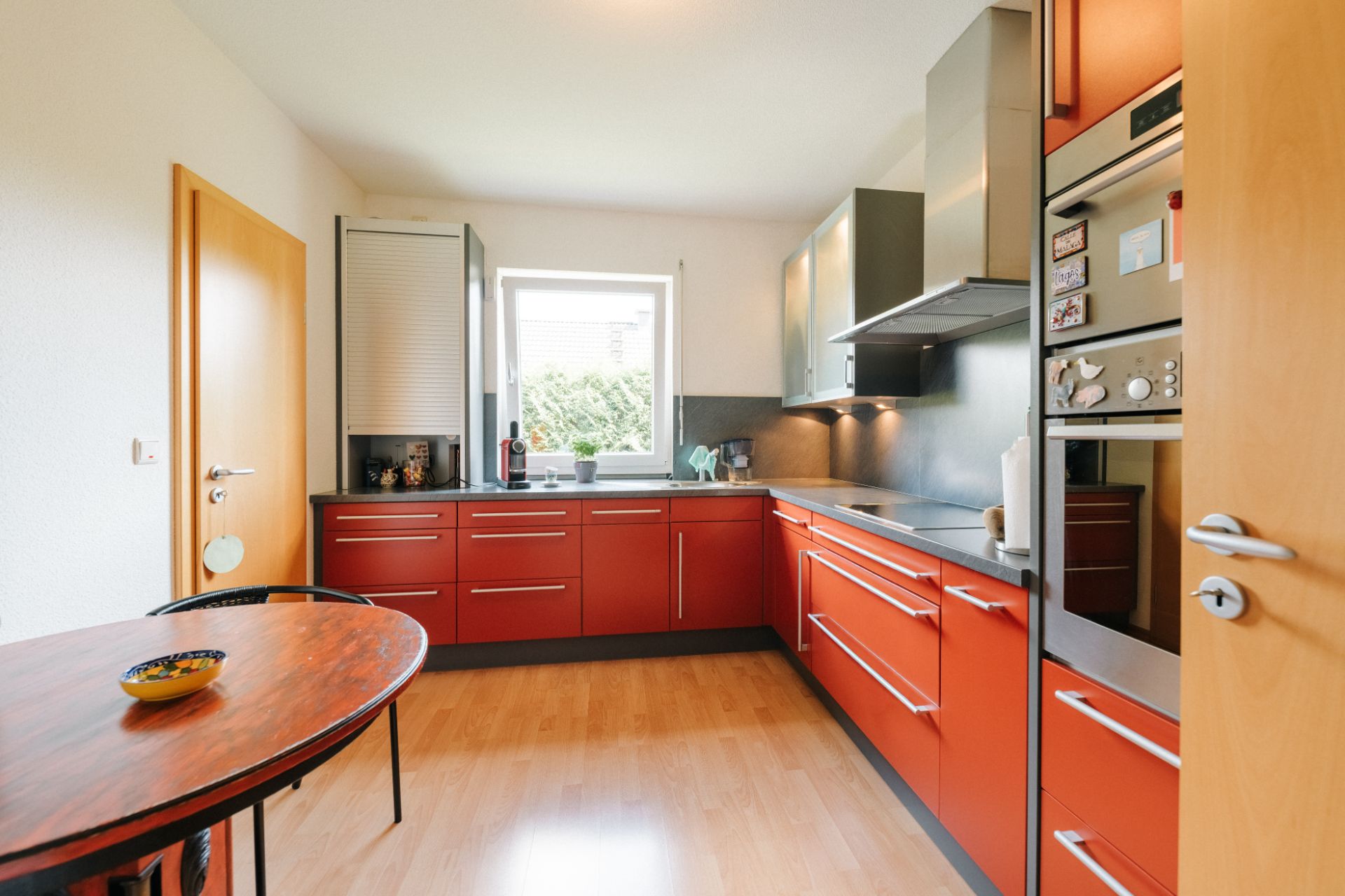 Moderne Küche mit roten Schränken, Holzfußboden, Tisch und Fenster im Hintergrund.