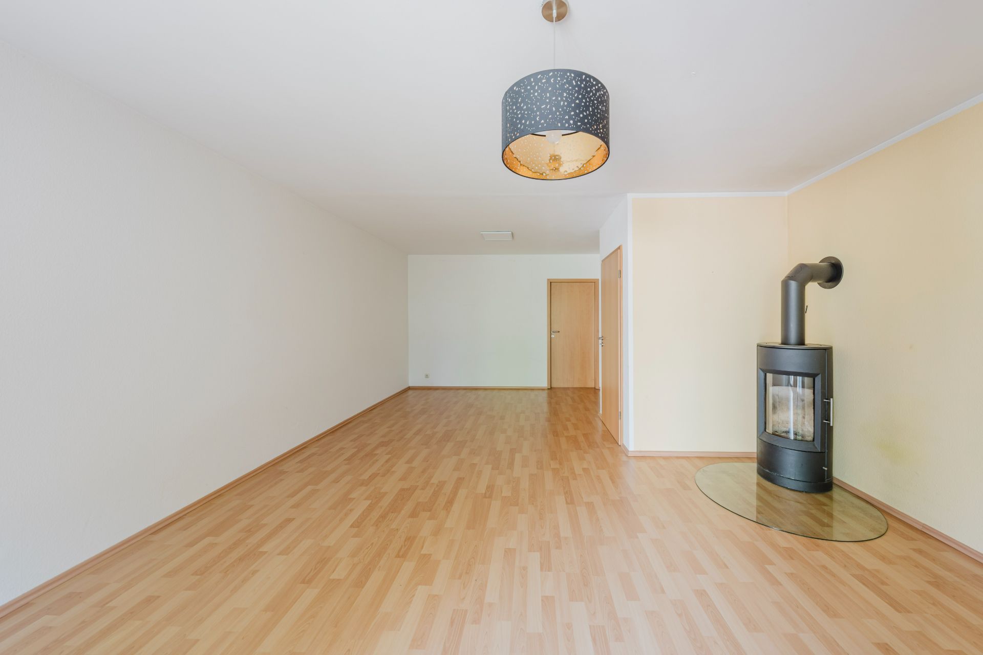 Leeres Wohnzimmer mit Holzfußboden, Kaminofen und Hängelampe an der Decke.