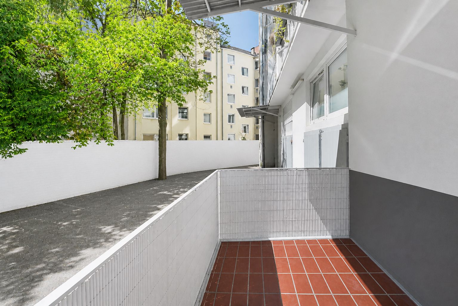 Kleine Terrasse mit roten Fliesen und weißem Zaun, Blick auf Bäume und Nachbargebäude.