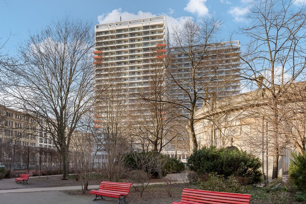 Wohnhochhaus hinter Bäumen mit roten Bänken im Park im Vordergrund.