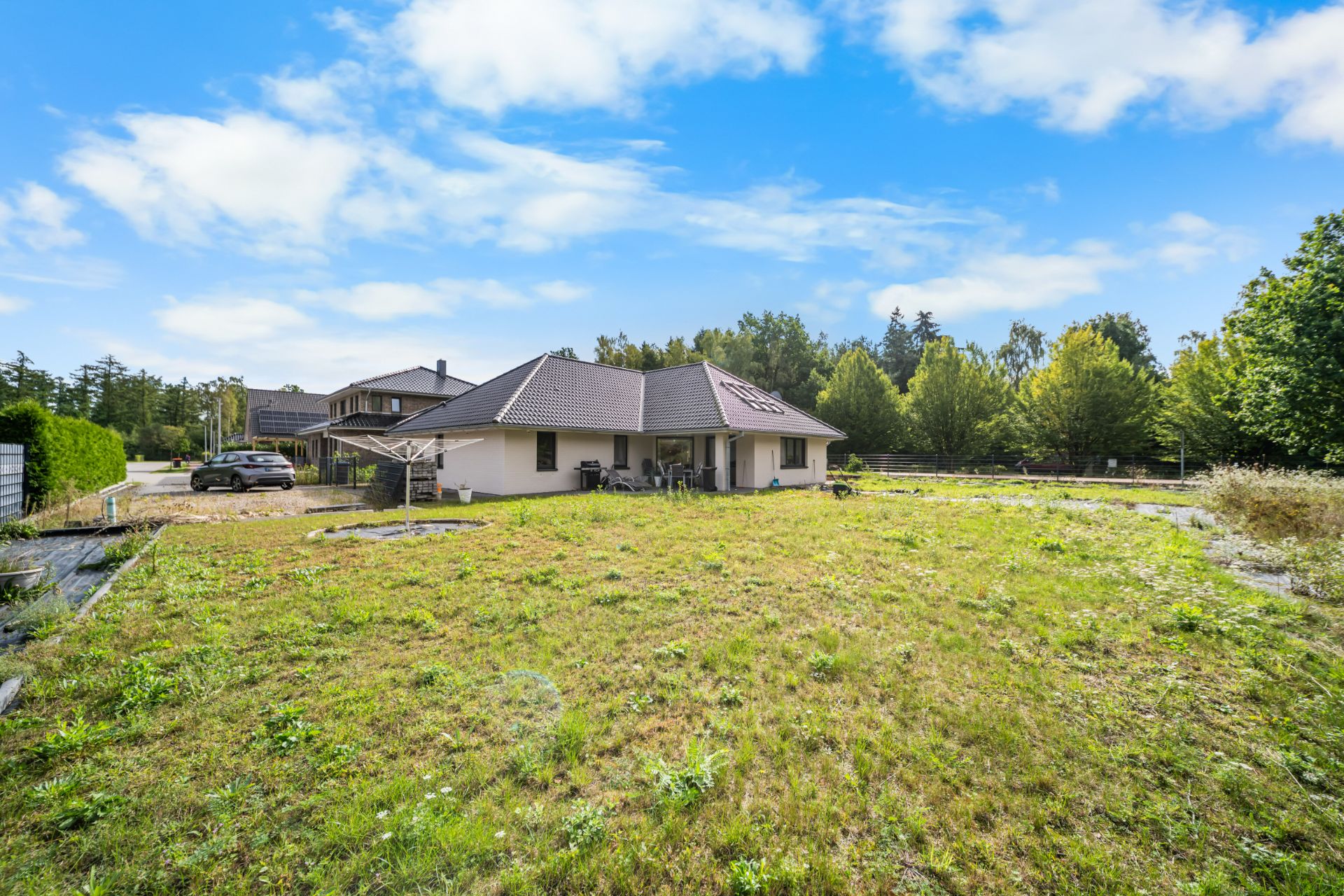 Einfamilienhaus mit Terrasse und großem Garten, umgeben von Bäumen unter blauem Himmel.