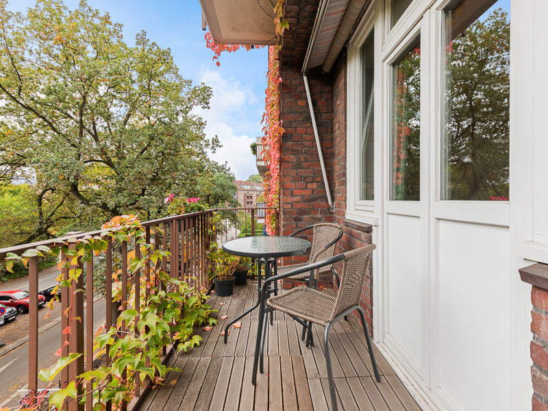 Balkon mit zwei Stühlen, rundem Tisch und Pflanzen am Geländer, Blick auf Bäume.