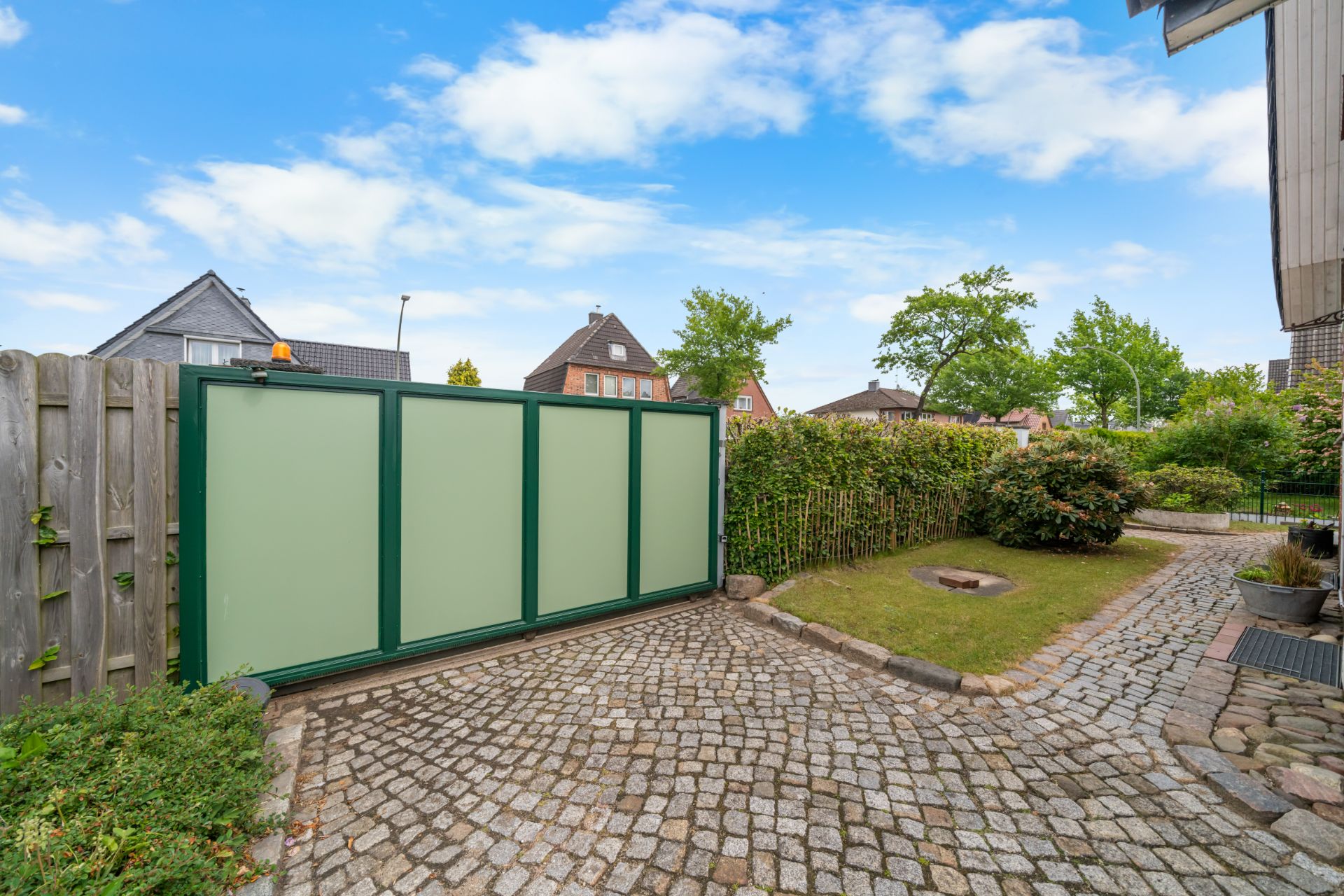 Gepflasterte Einfahrt mit grünem Tor, Garten und einigen Büschen im Wohngebiet.