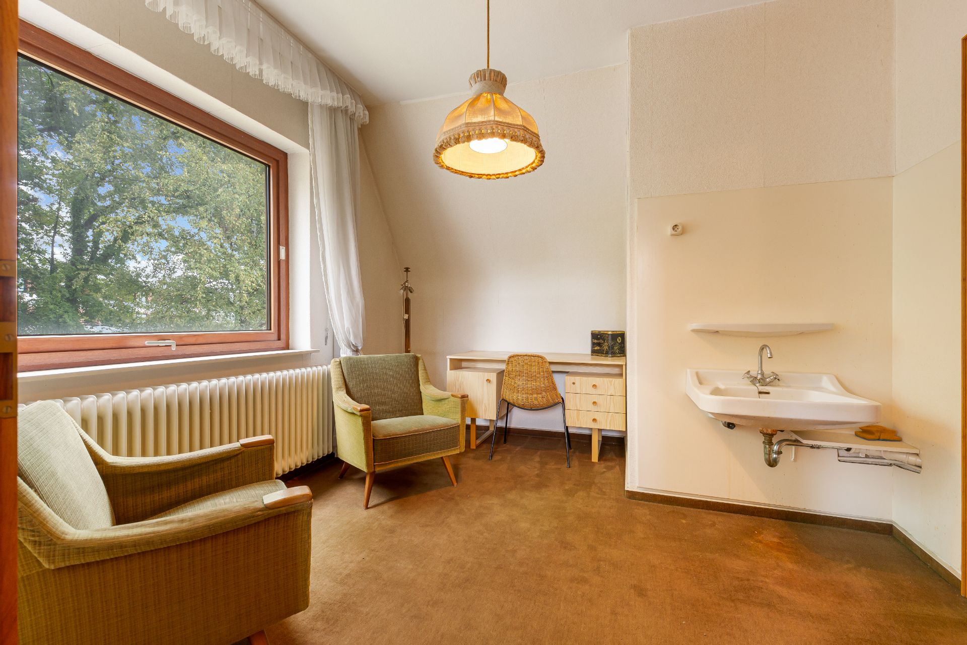 Zimmer mit zwei Sesseln, großem Fenster, Schreibtisch und Waschbecken an der Wand.