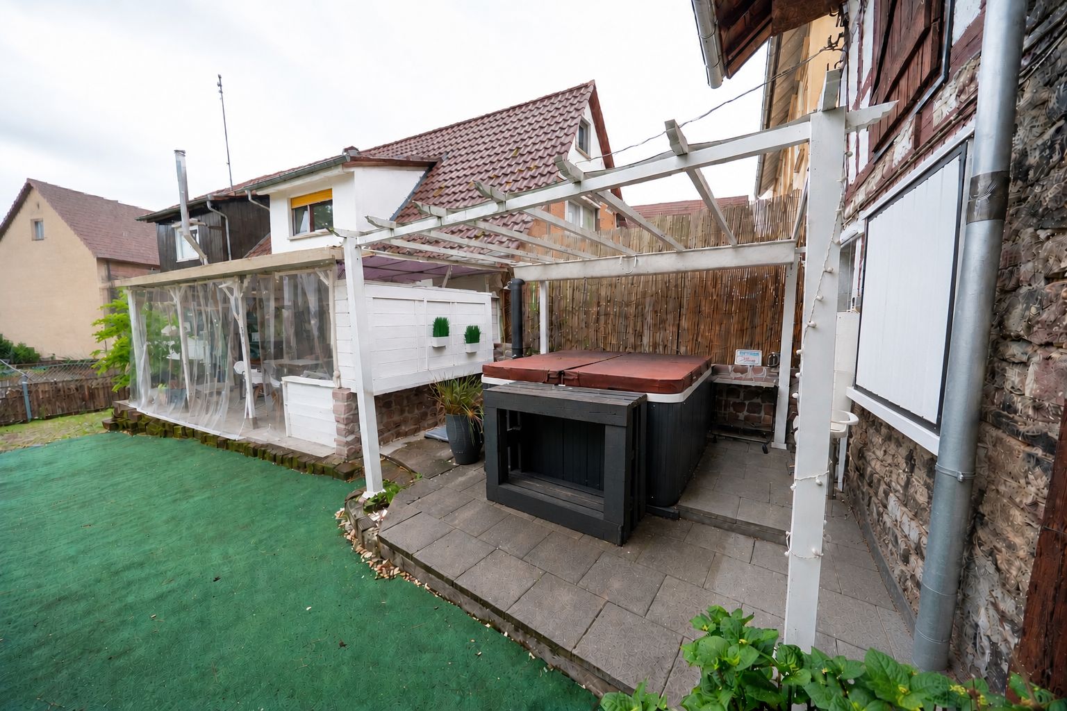 Gartenbereich mit überdachtem Whirlpool und Kunstrasen vor einem Wohnhaus.