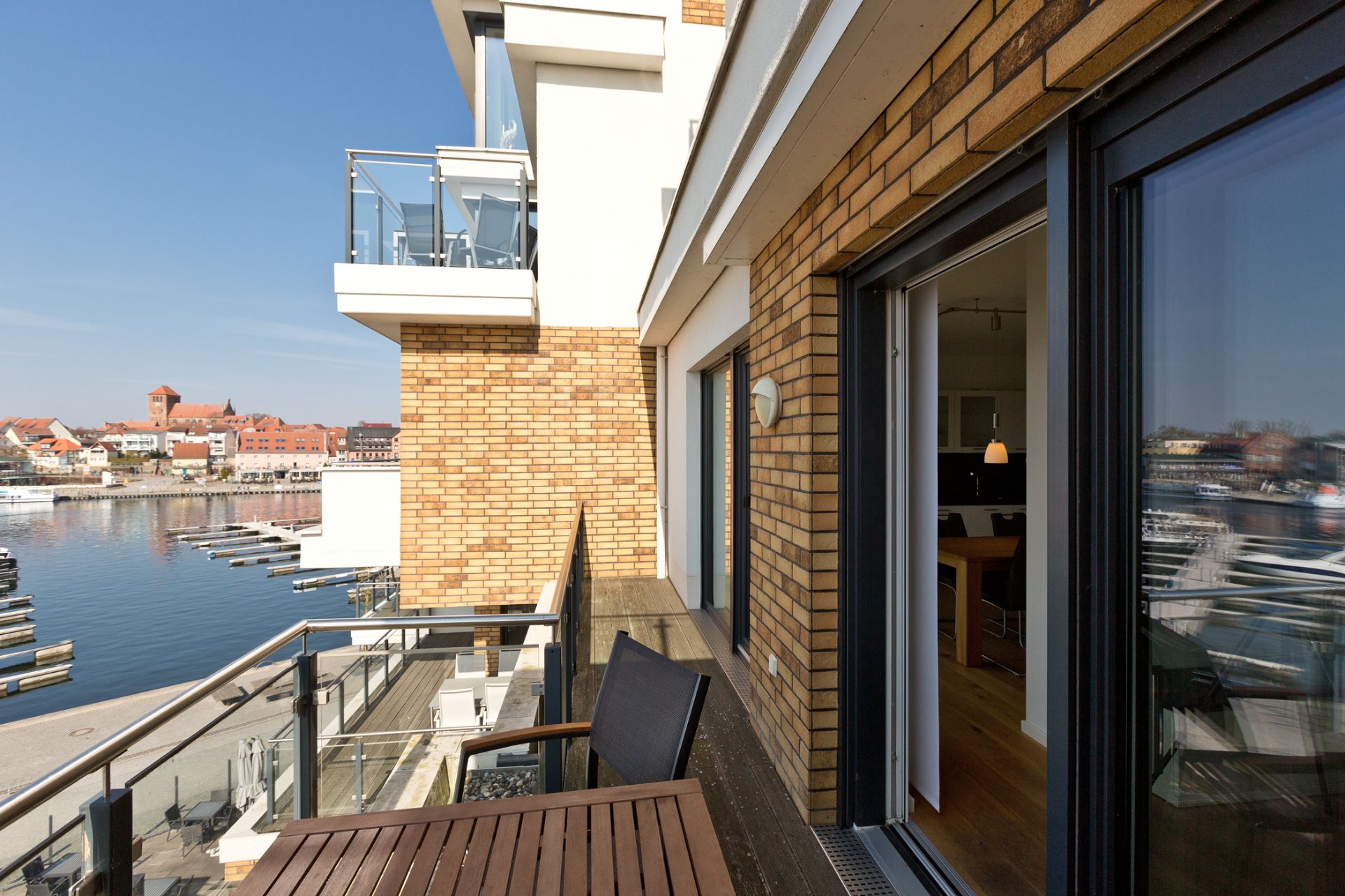 Balkon mit Holztisch und Stühlen, Blick auf einen Hafen und Backsteinfassade.