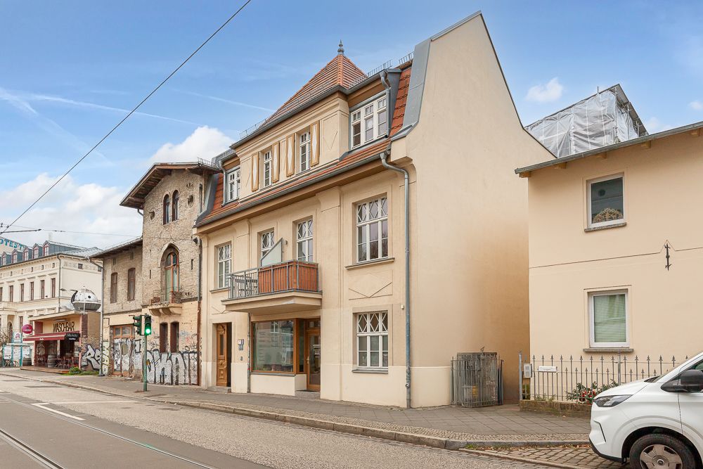 Dreistöckiges Wohnhaus mit Balkon an einer Stadtstraße, daneben weitere Gebäude.