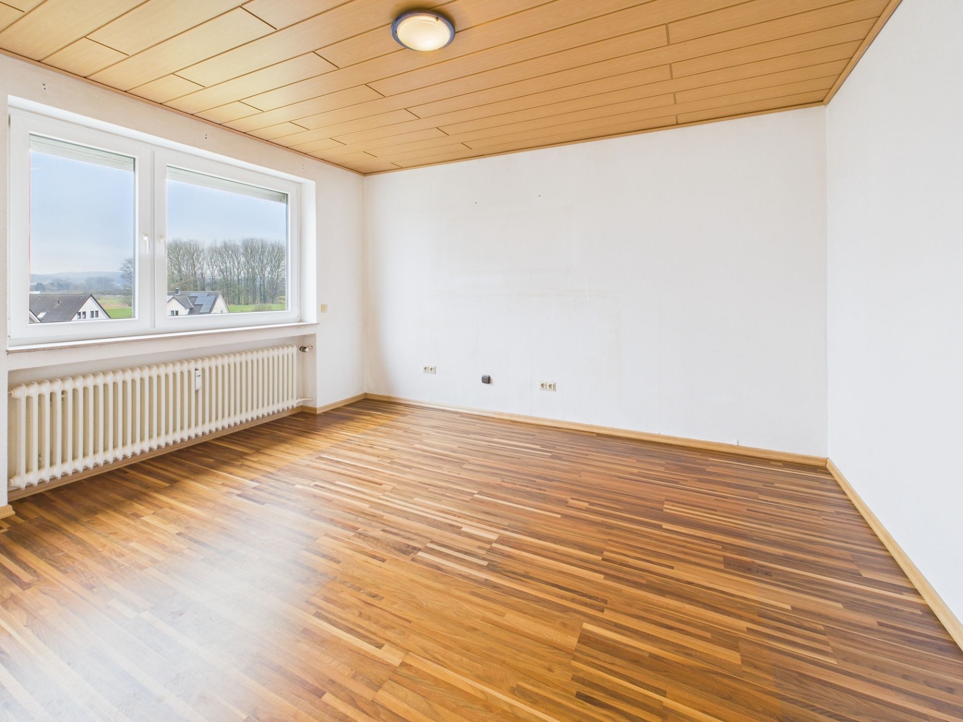 Leerer Raum mit Holzfußboden, weißer Wand, großem Fenster und Heizkörper.
