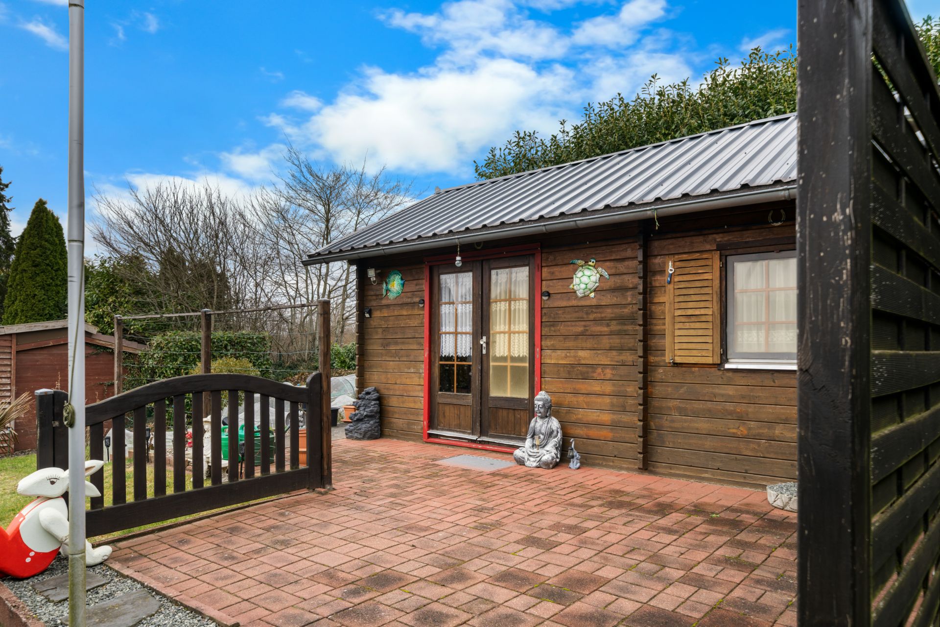 Kleines Holzhaus mit Terrasse, Buddha-Statue und Gartenzaun bei bewölktem Himmel.