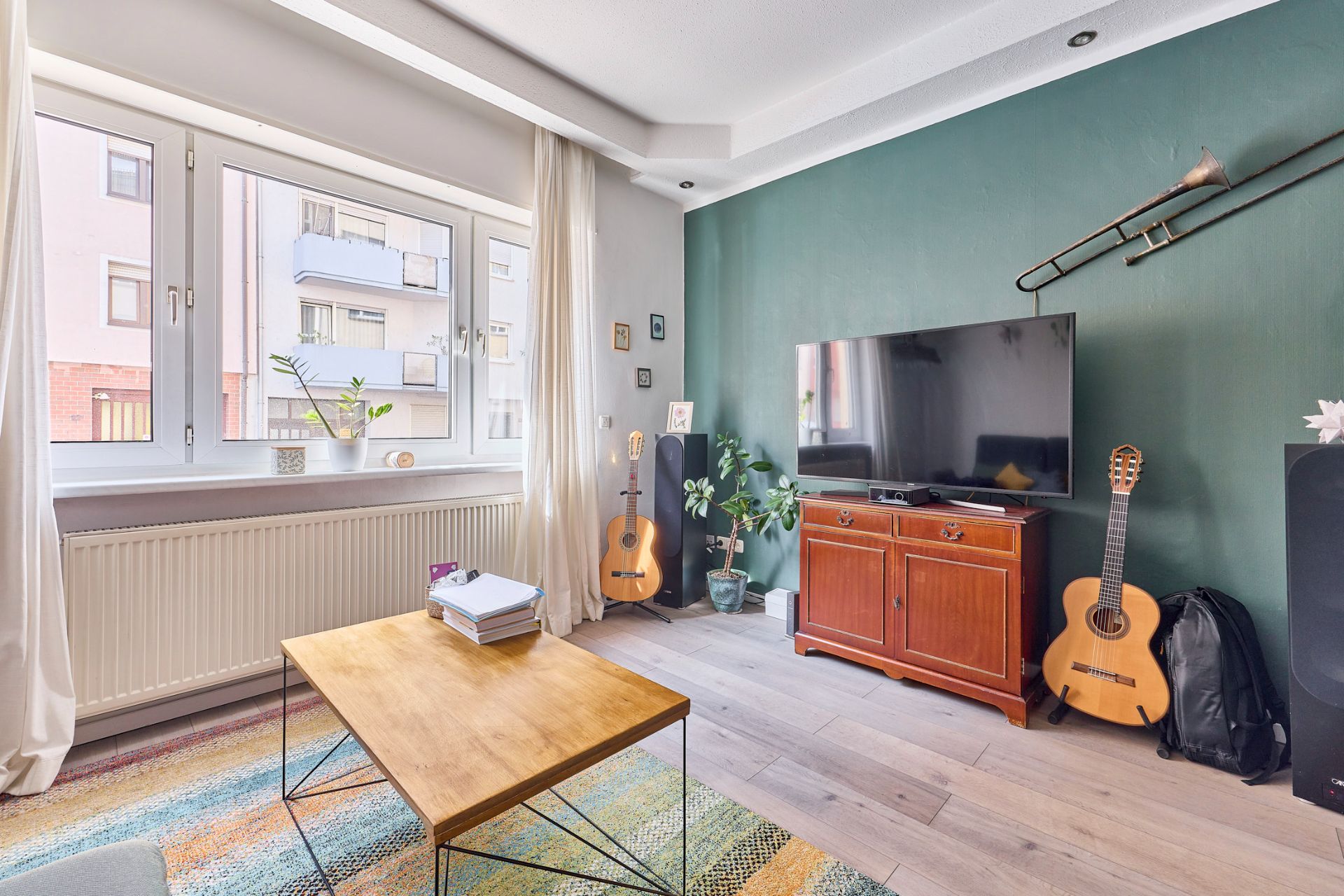 Helles Wohnzimmer mit grün gestrichener Wand, Fernseher, zwei Gitarren und großem Fenster.