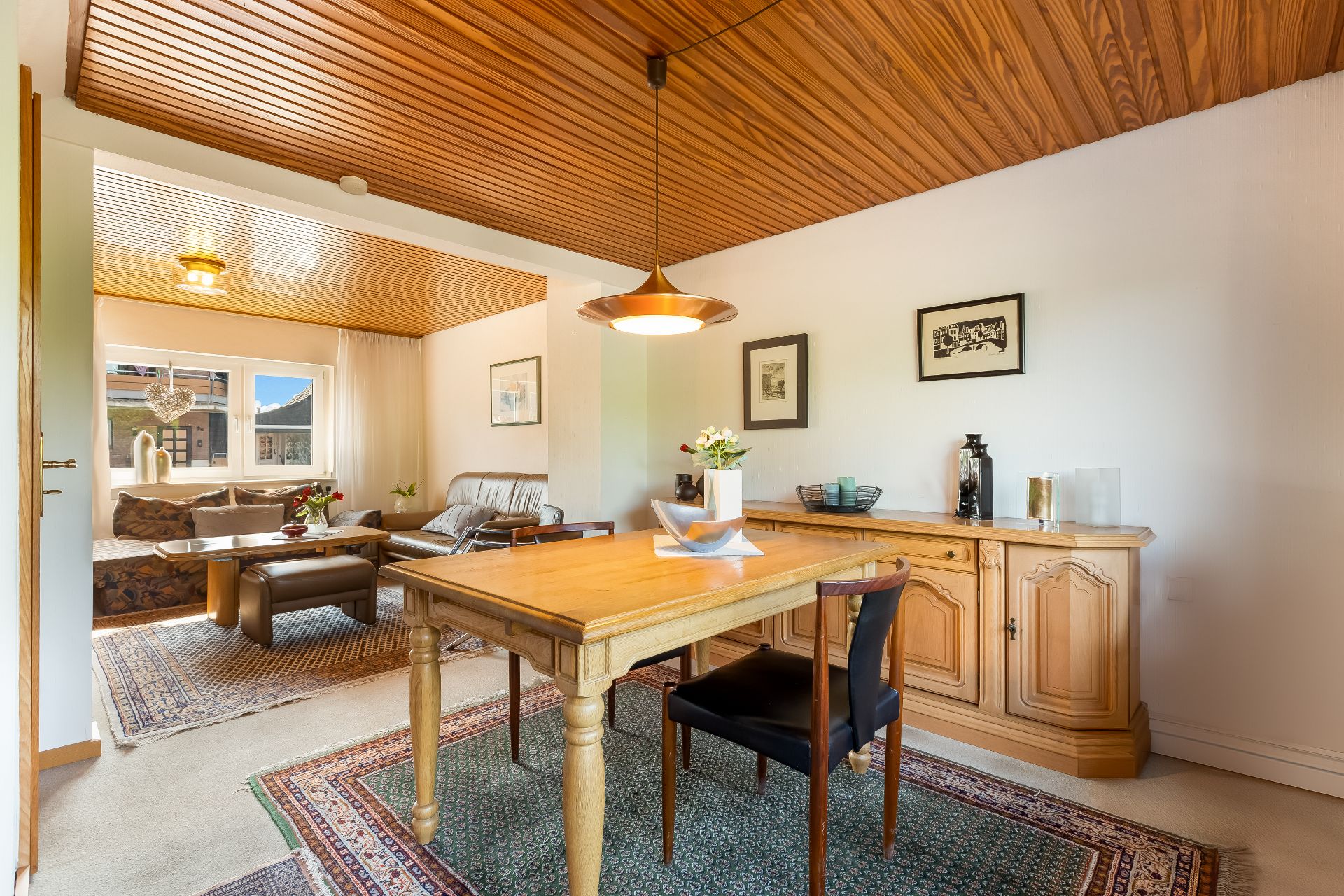 Helles Esszimmer mit Holztisch, Teppichen und angrenzendem Wohnzimmer mit Sofa und Fenster.