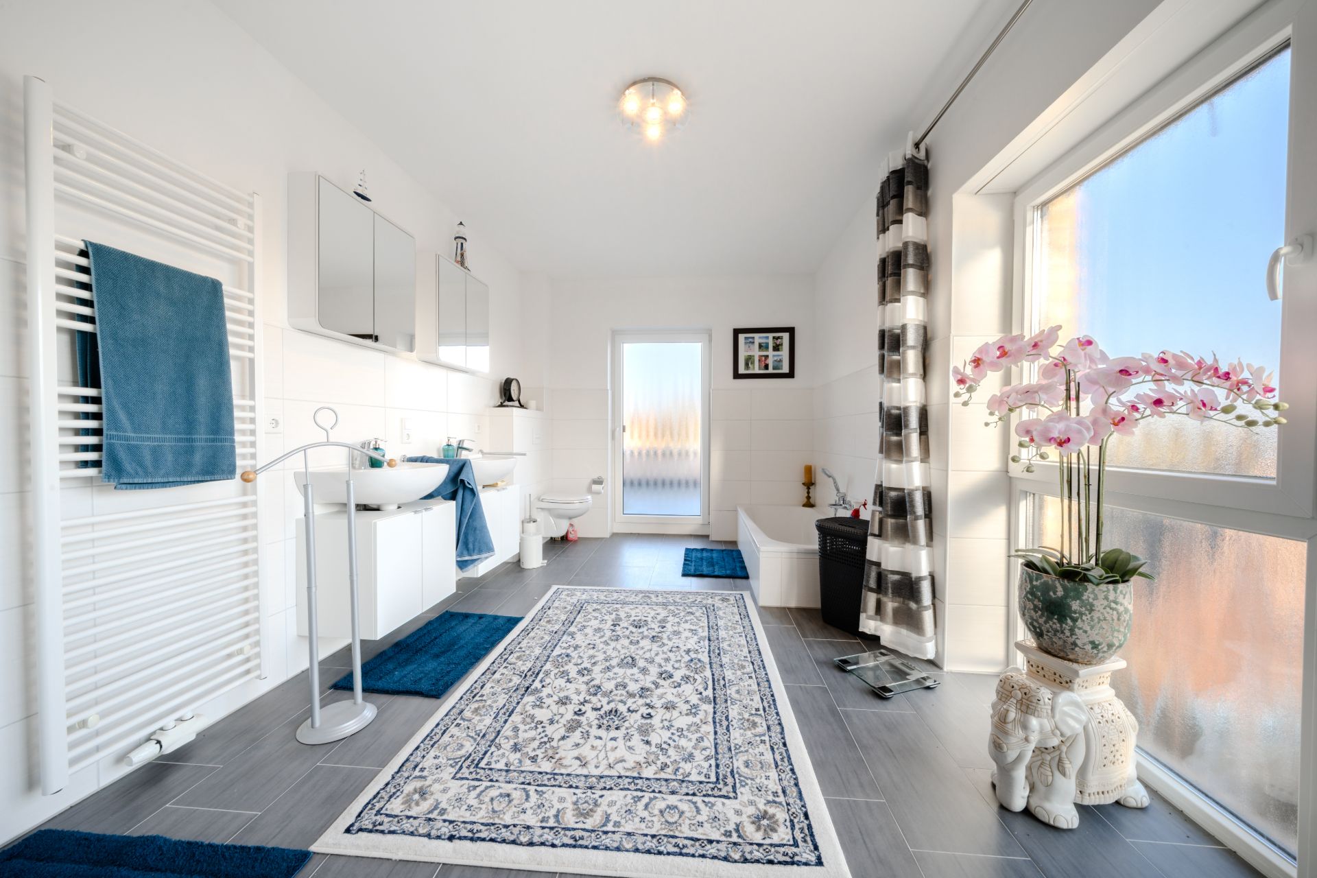 Helles Badezimmer mit Teppich, Badewanne, Waschbecken, großem Fenster und Pflanzen.