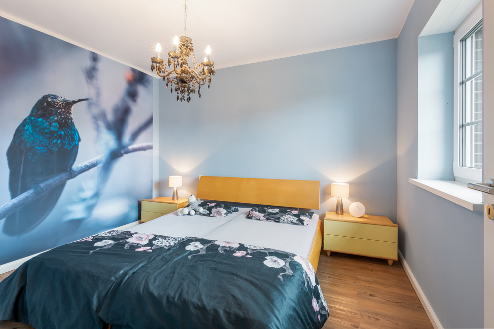 Schlafzimmer mit Doppelbett, blauer Wand, Vogelbild, Kronleuchter und zwei Nachttischen mit Lampen.