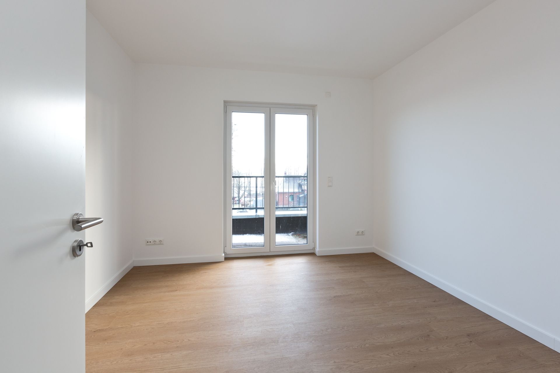 Leerer Raum mit Holzboden, weißen Wänden und einer Glastür zu einem Balkon.