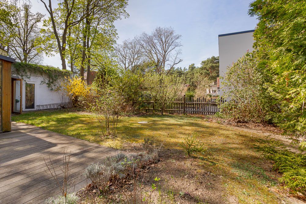 Garten mit Terrasse, Büschen und Bäumen, Holzzaun im Hintergrund.