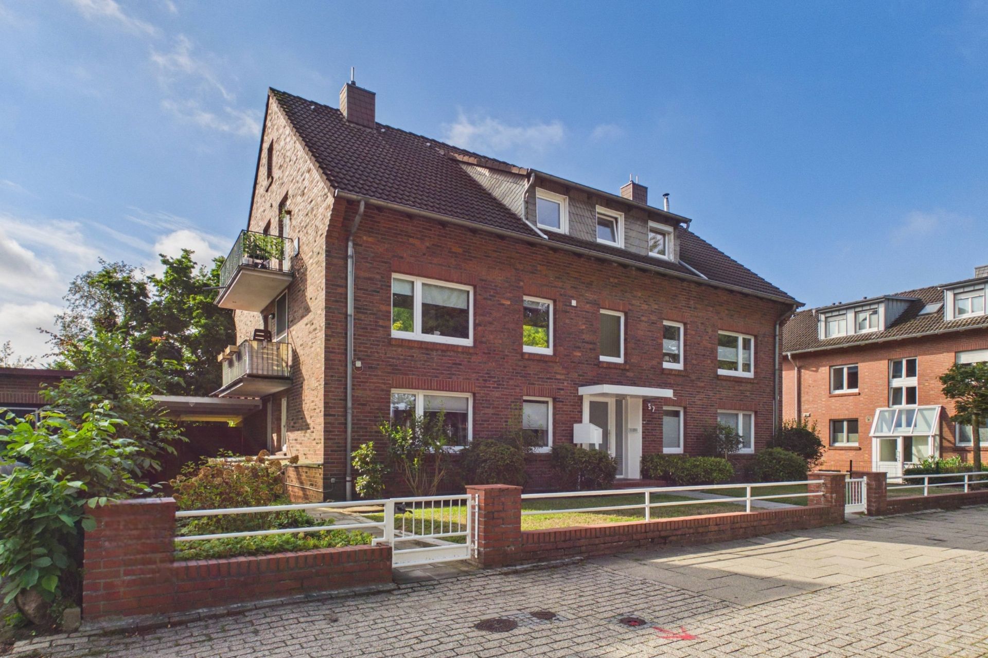 Dreistöckiges Wohnhaus aus rotem Backstein mit Vorgarten und weißem Zaun an einer ruhigen Straße.