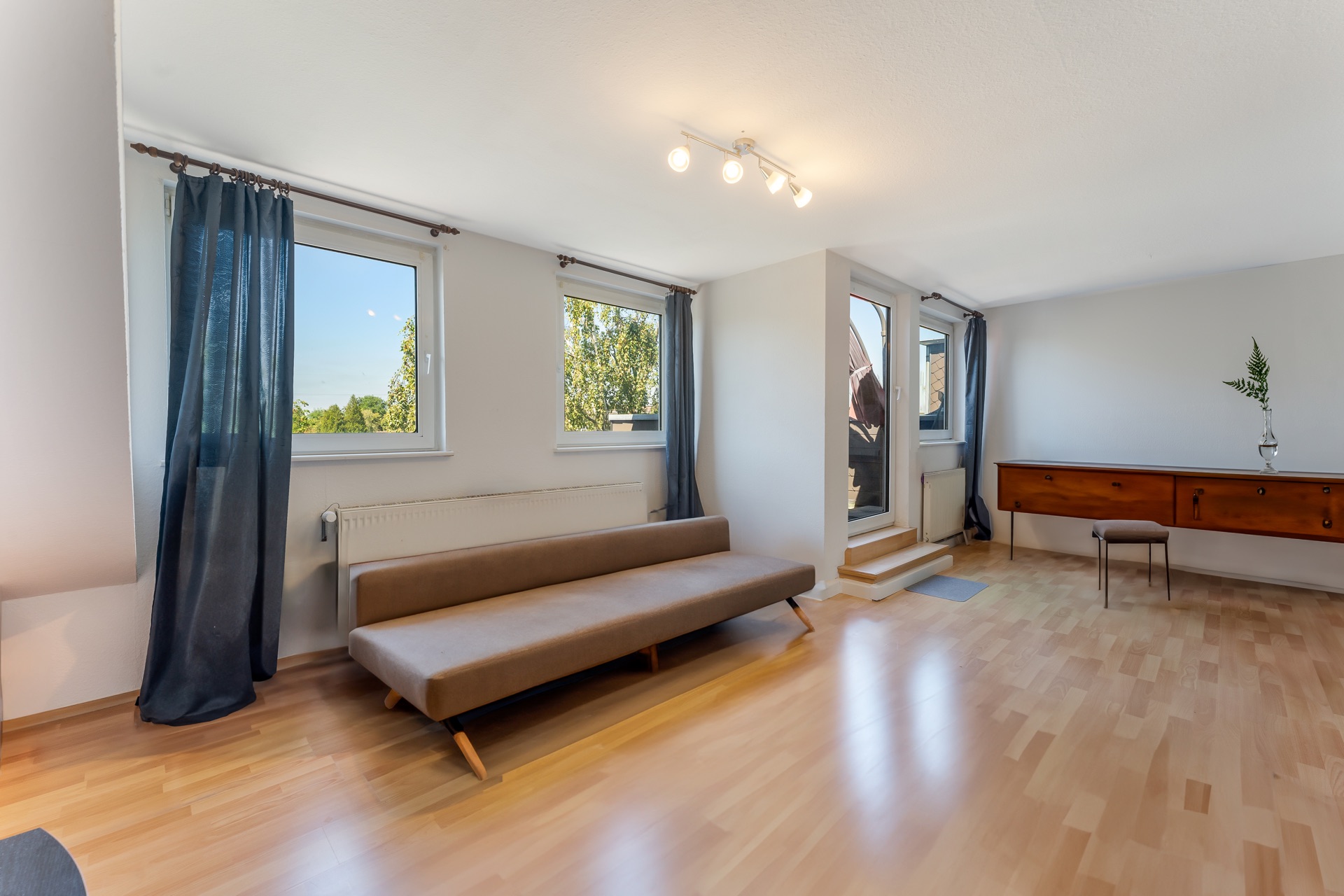 Helles Wohnzimmer mit braunem Sofa, Holzkommode, zwei Fenstern und Tür zum Balkon.