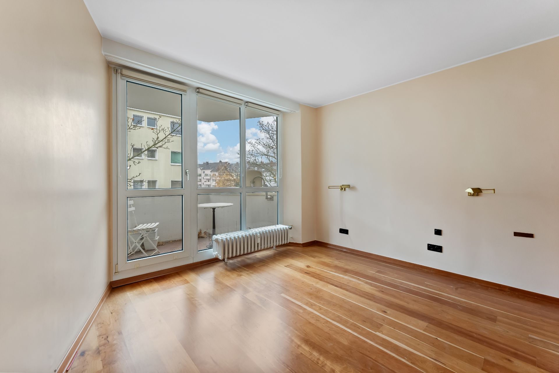 Leeres Zimmer mit Holzboden, großen Fenstern und Zugang zu einem kleinen Balkon mit Stuhl und Tisch.