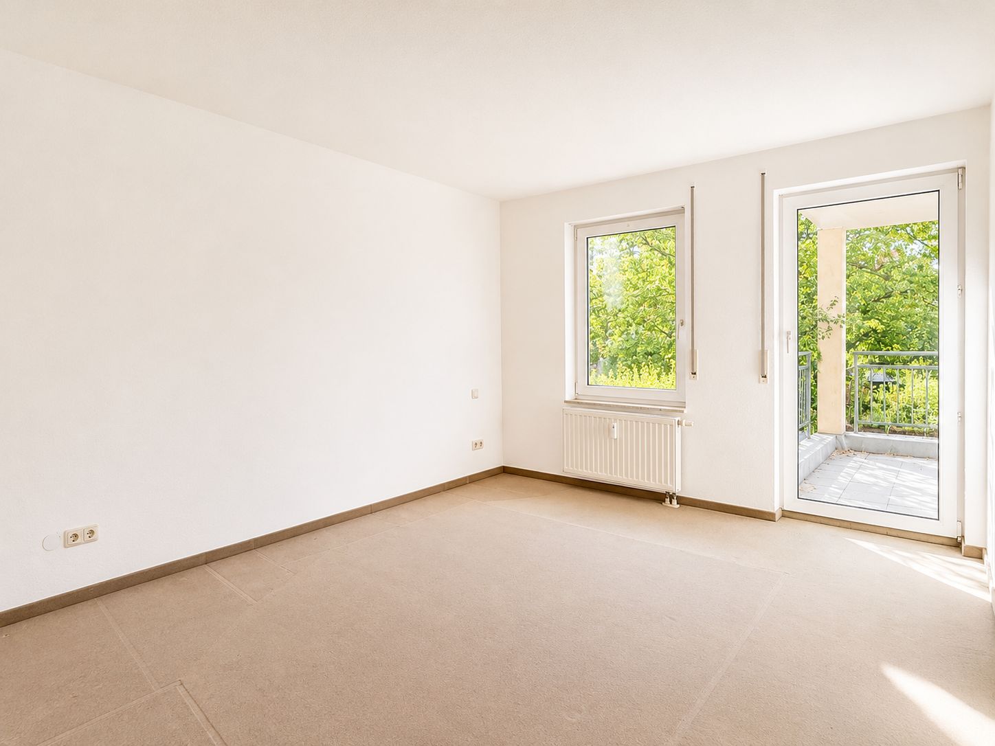 Leeres Zimmer mit Teppichboden, Fenster und Tür zum Balkon, viel Tageslicht.
