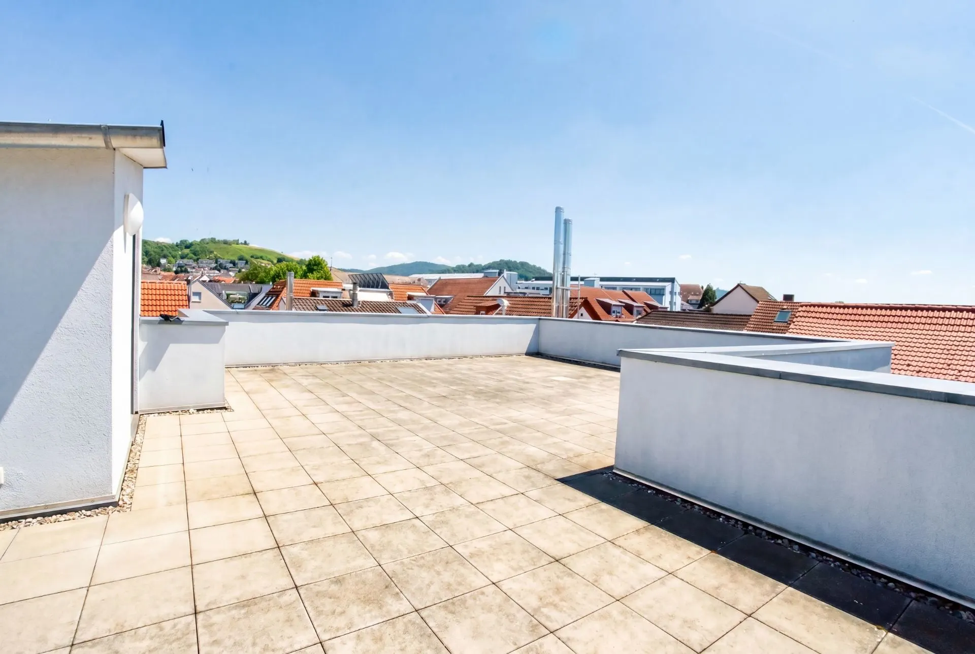 Große, geflieste Dachterrasse mit Blick auf Dächer und Hügel im Hintergrund.