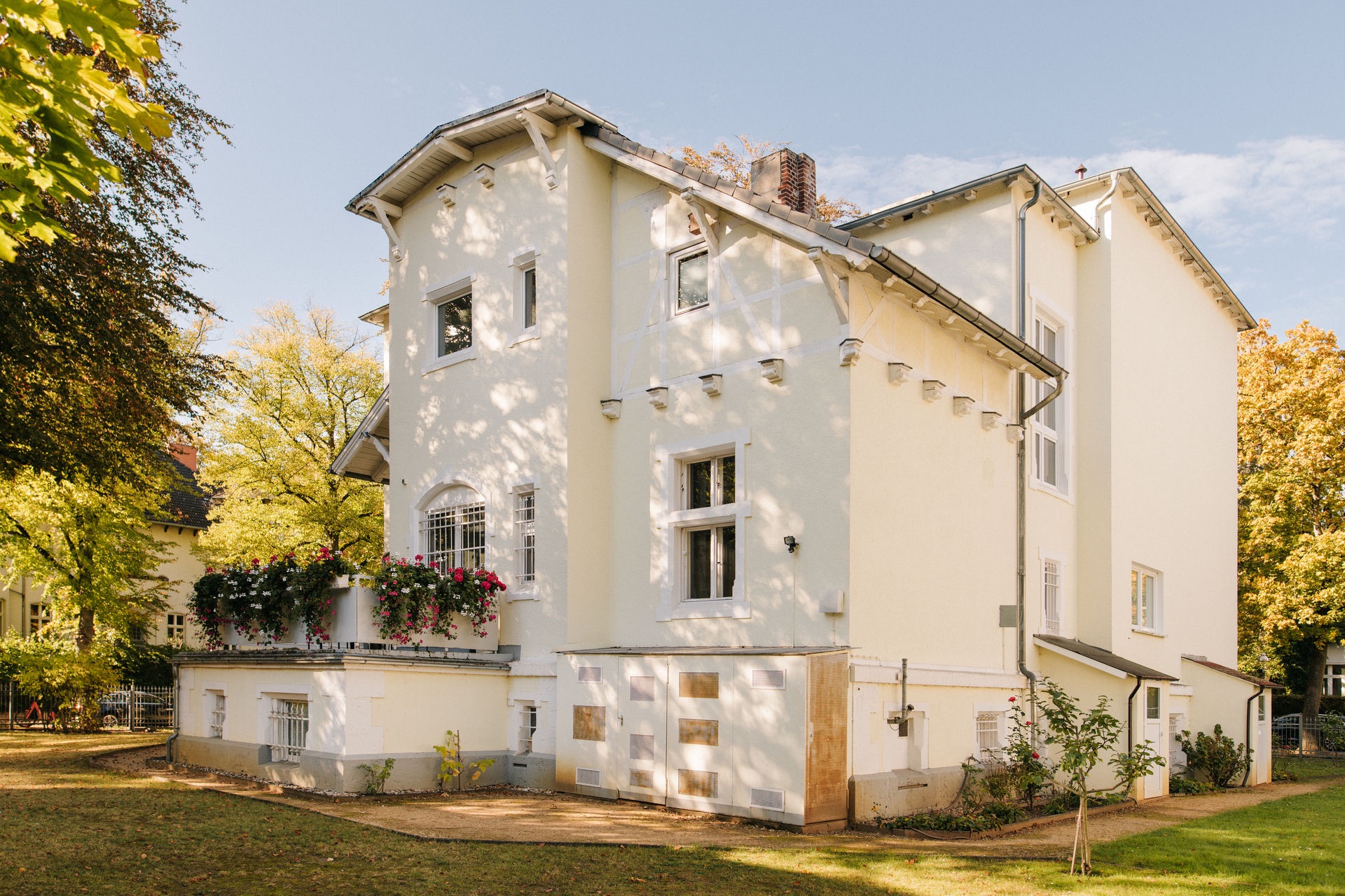 Helles, zweistöckiges Wohnhaus mit Blumen auf dem Balkon und umgeben von Bäumen.