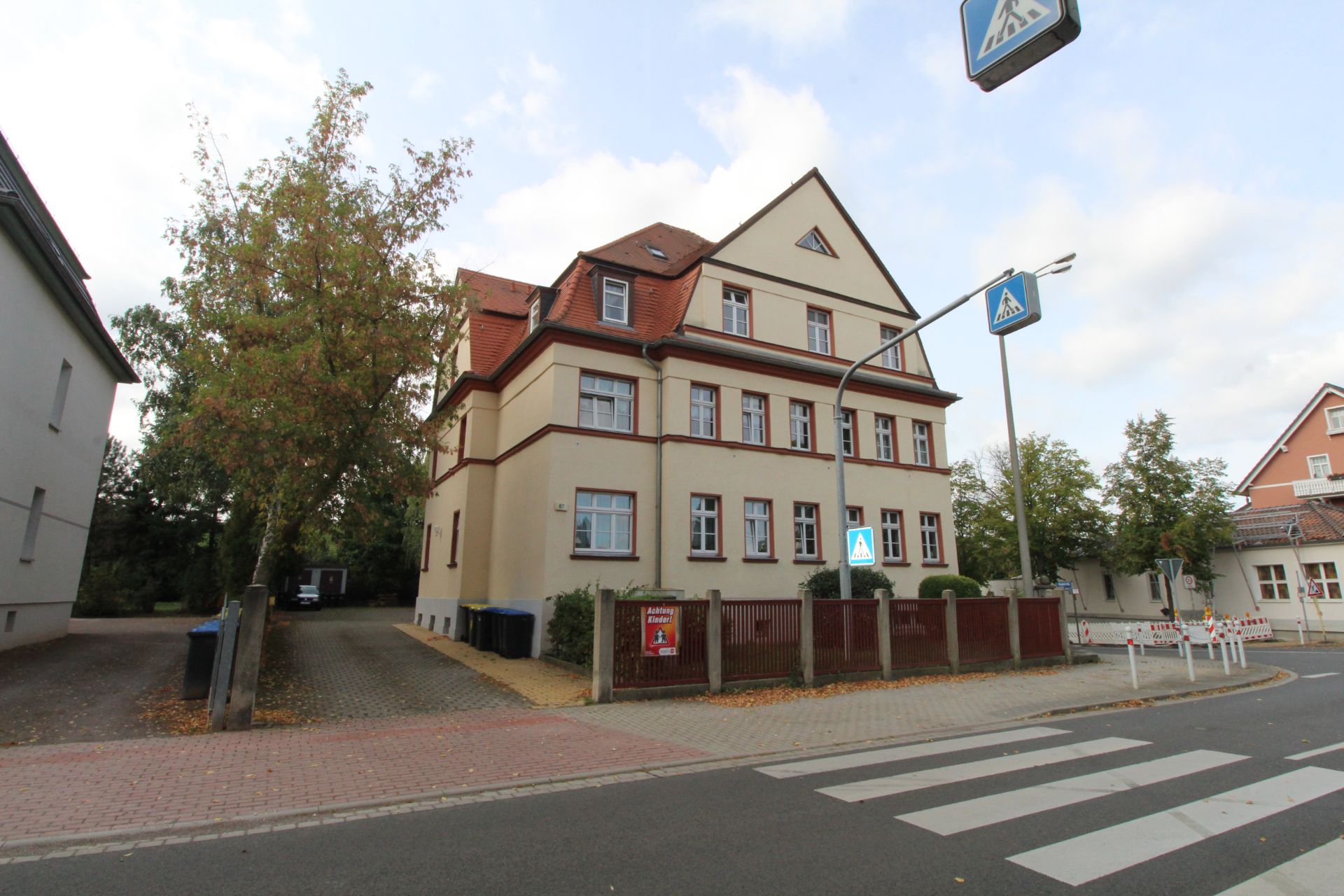 Wohnhaus mit rotem Dach an einer Straße mit Zebrastreifen und Zäunen im Vordergrund.