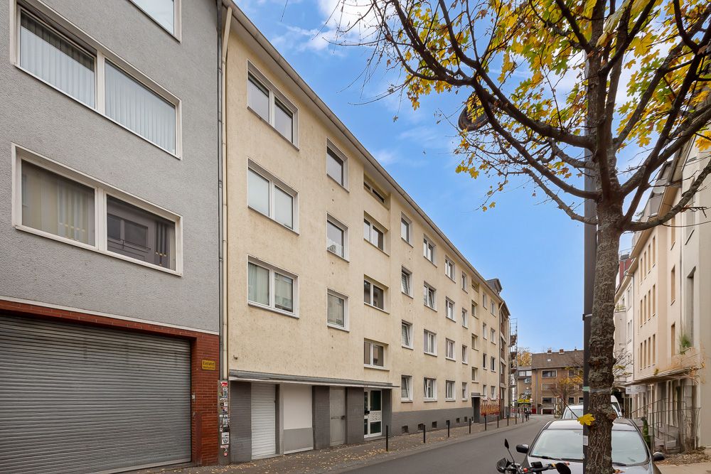 Wohnhaus mit mehreren Stockwerken an einer ruhigen Straße, mit Bäumen und geparkten Autos.