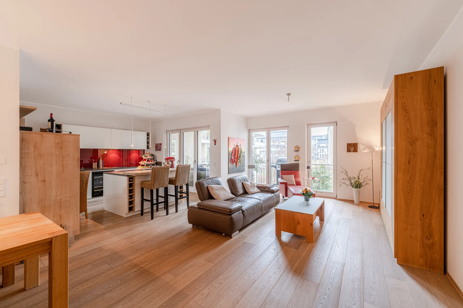 Helles Wohnzimmer mit offener Küche, Holzboden, Sofa, Esstisch und großen Fenstern.