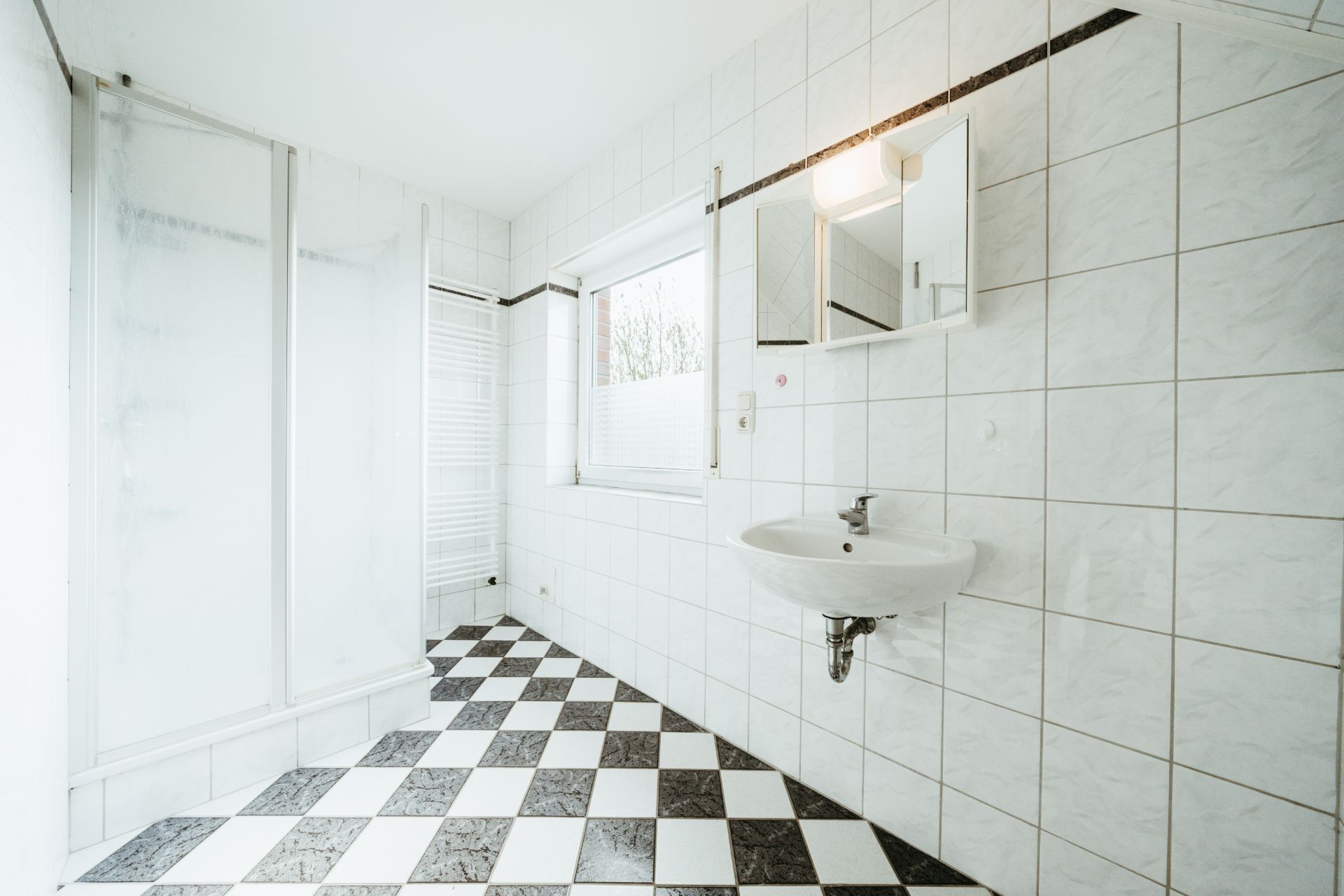 Helles Badezimmer mit karierten Fliesen, Dusche, Fenster, Waschbecken und Spiegel.