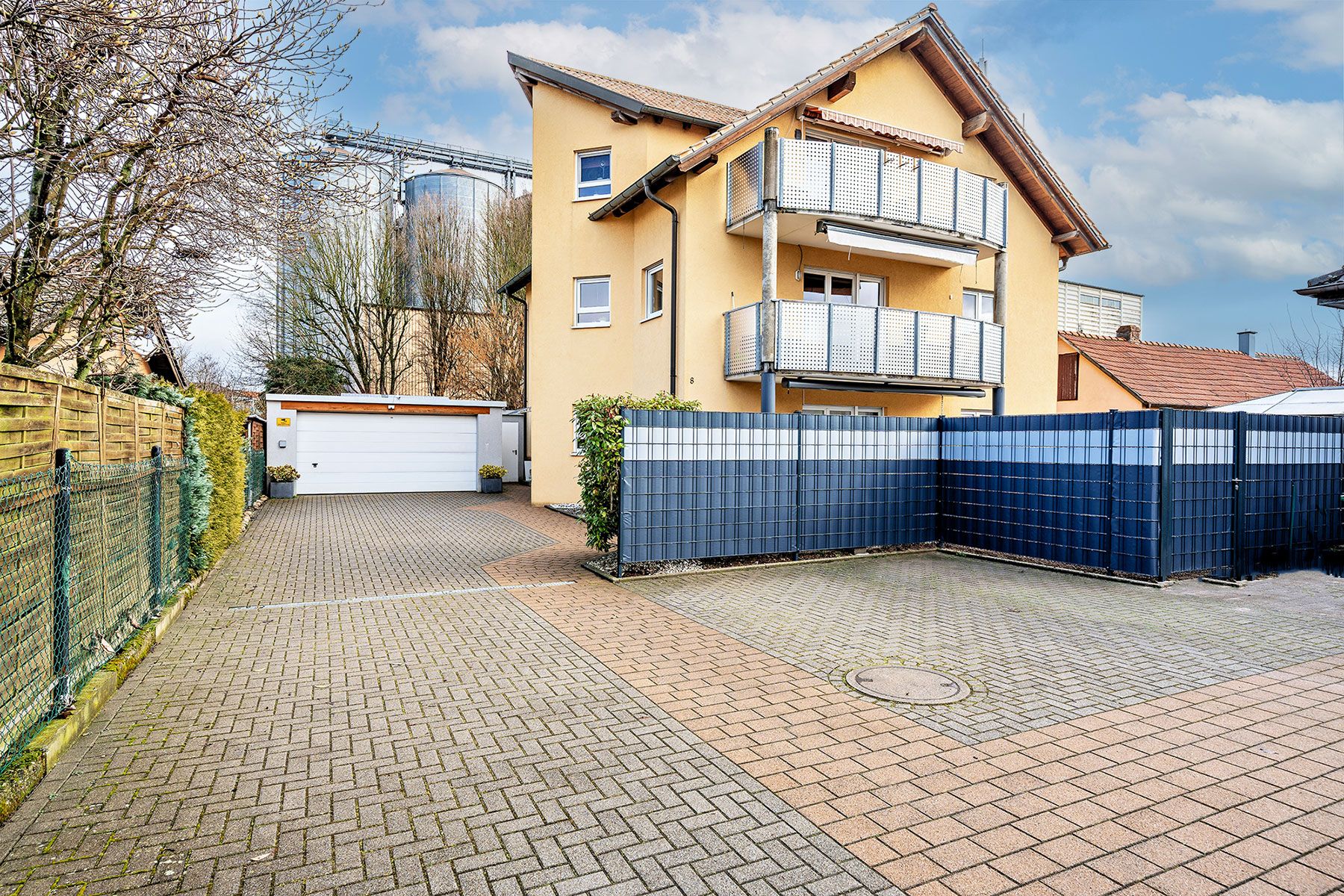 Modernes gelbes Wohnhaus mit zwei Balkonen, Einfahrt und Garage, umgeben von Zaun und Pflanzen.