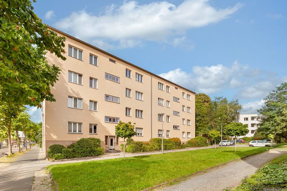 Mehrstöckiges Wohngebäude mit beige Fassade und mehreren Fenstern, umgeben von Grünflächen und Gehweg.