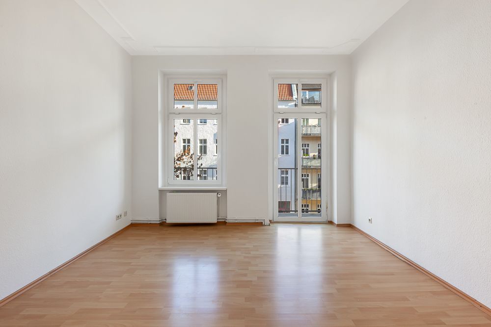 Heller leerer Raum mit Parkettboden, zwei Fenstern und einer Glastür zu einem Balkon.