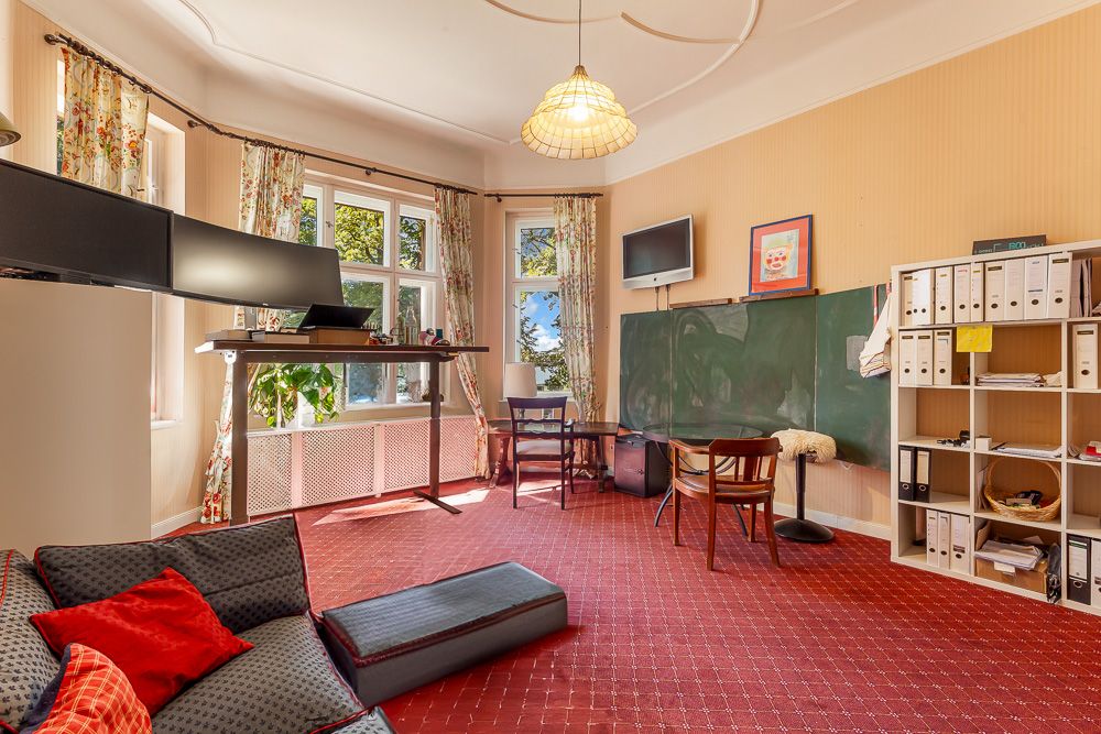 Helles Zimmer mit Schreibtisch, großen Monitoren, Tafel, Regal und Sesseln auf rotem Teppich.