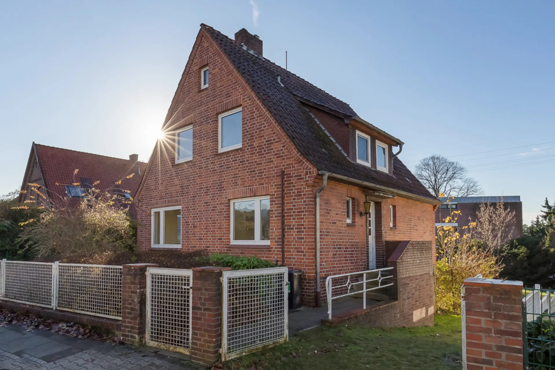 Backsteinhaus mit Satteldach, weißen Fenstern, Vorgarten und Metallzaun bei Sonnenschein.