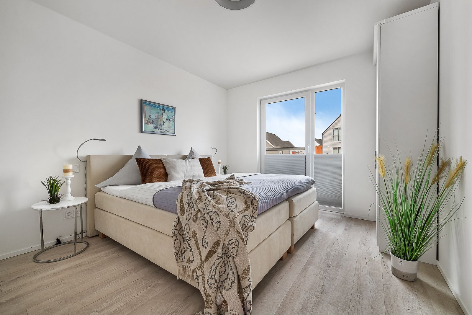 Helles Schlafzimmer mit Doppelbett, Nachttisch, Bild an der Wand und Zugang zum Balkon.