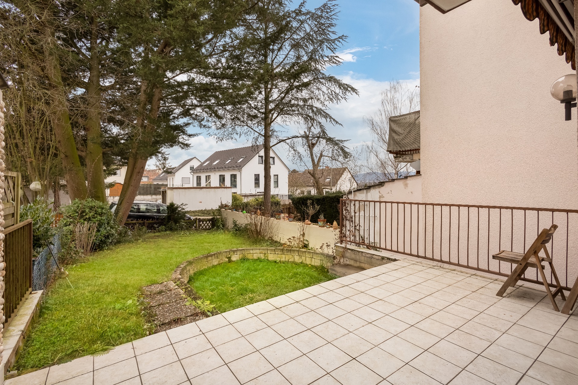 Geflieste Terrasse mit Stuhl, angrenzender Garten und Bäumen, Häuser im Hintergrund.
