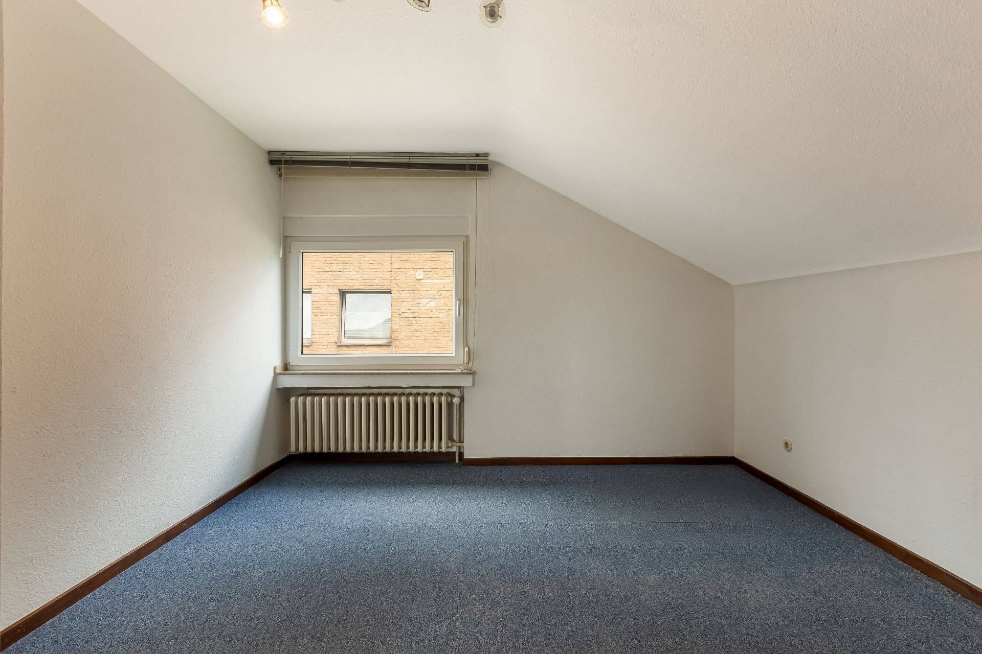 Leeres Zimmer mit blauem Teppich, schräger Decke und Fenster mit Heizkörper darunter.