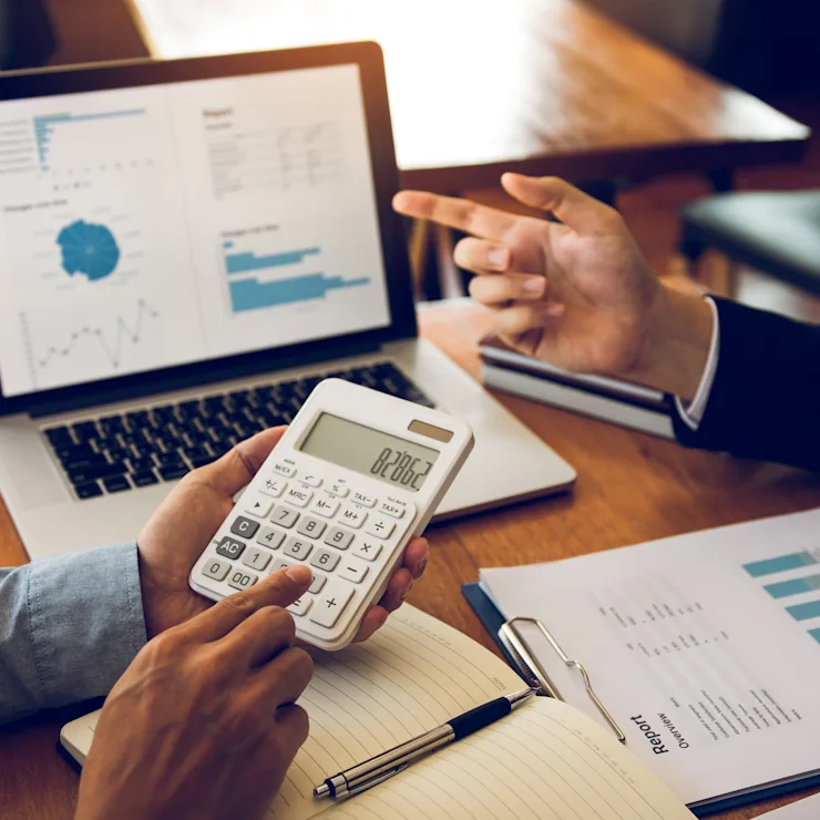 Zwei Personen besprechen Diagramme auf einem Laptop, mit Taschenrechner und Berichten auf dem Tisch.