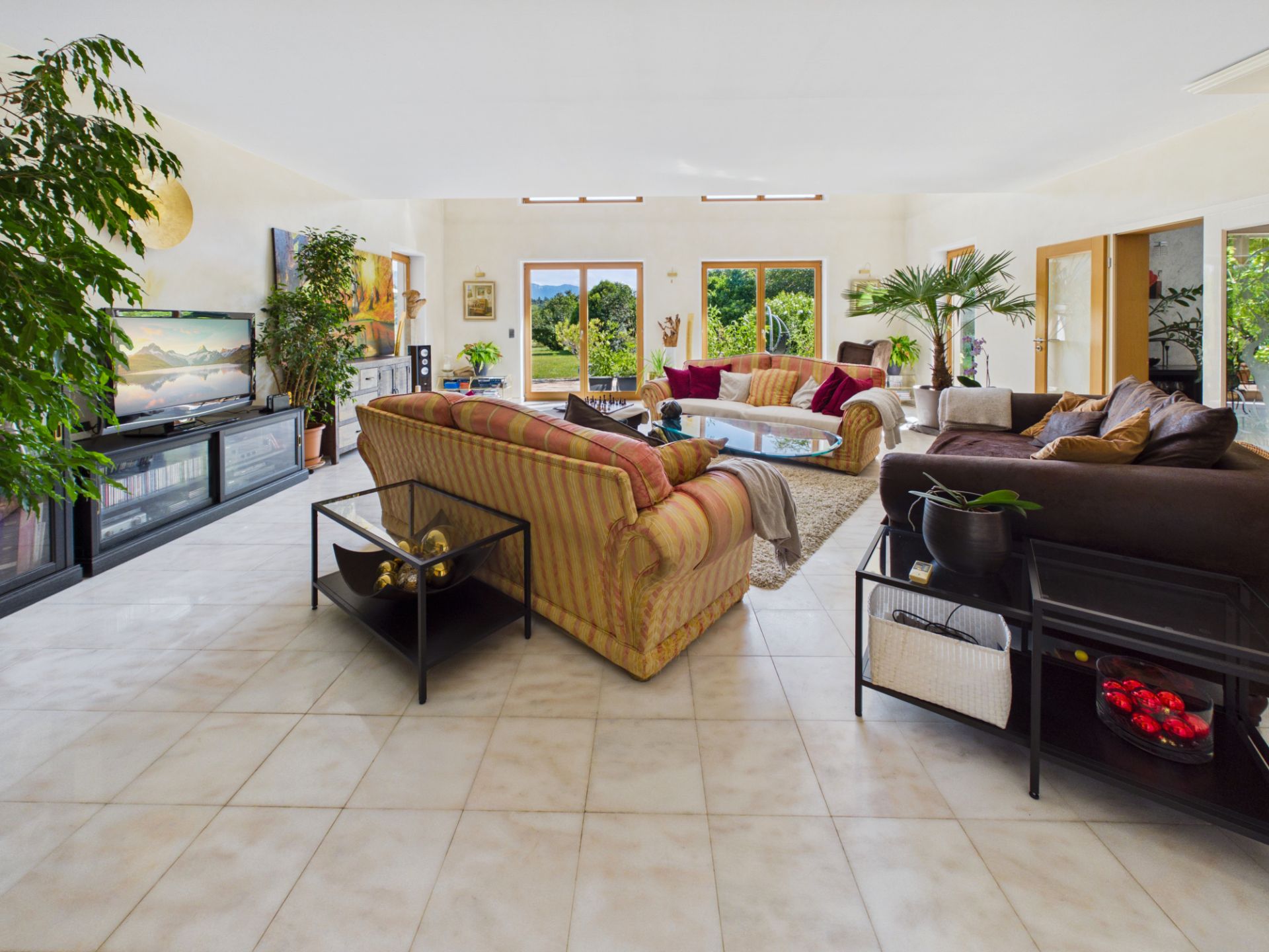 Großes Wohnzimmer mit mehreren Sofas, Pflanzen, Couchtisch und großen Fenstern mit Gartenblick.