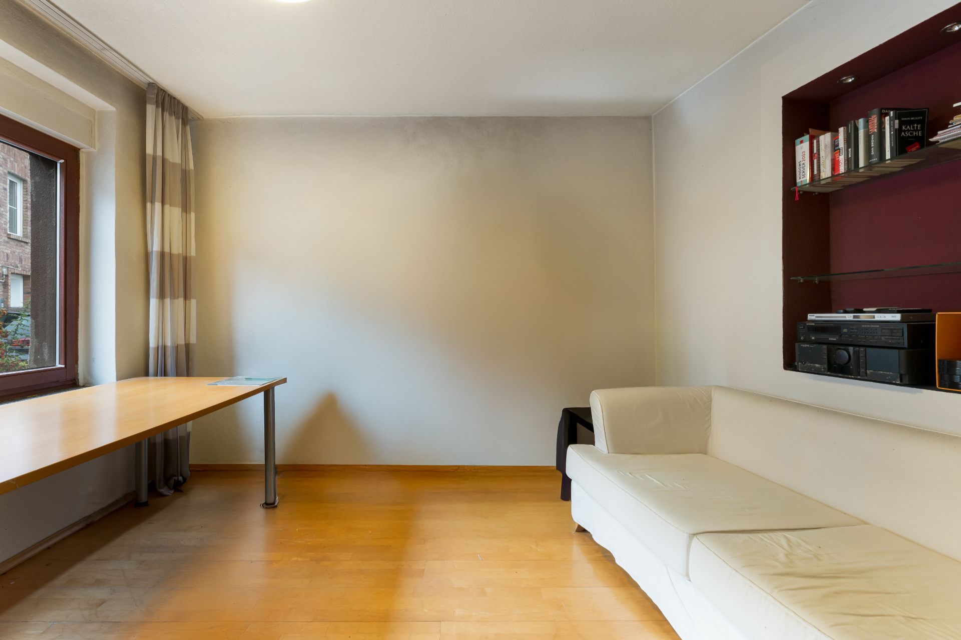 Helles Zimmer mit Ecksofa, Holzboden, großem Schreibtisch am Fenster und Wandregal mit Büchern.