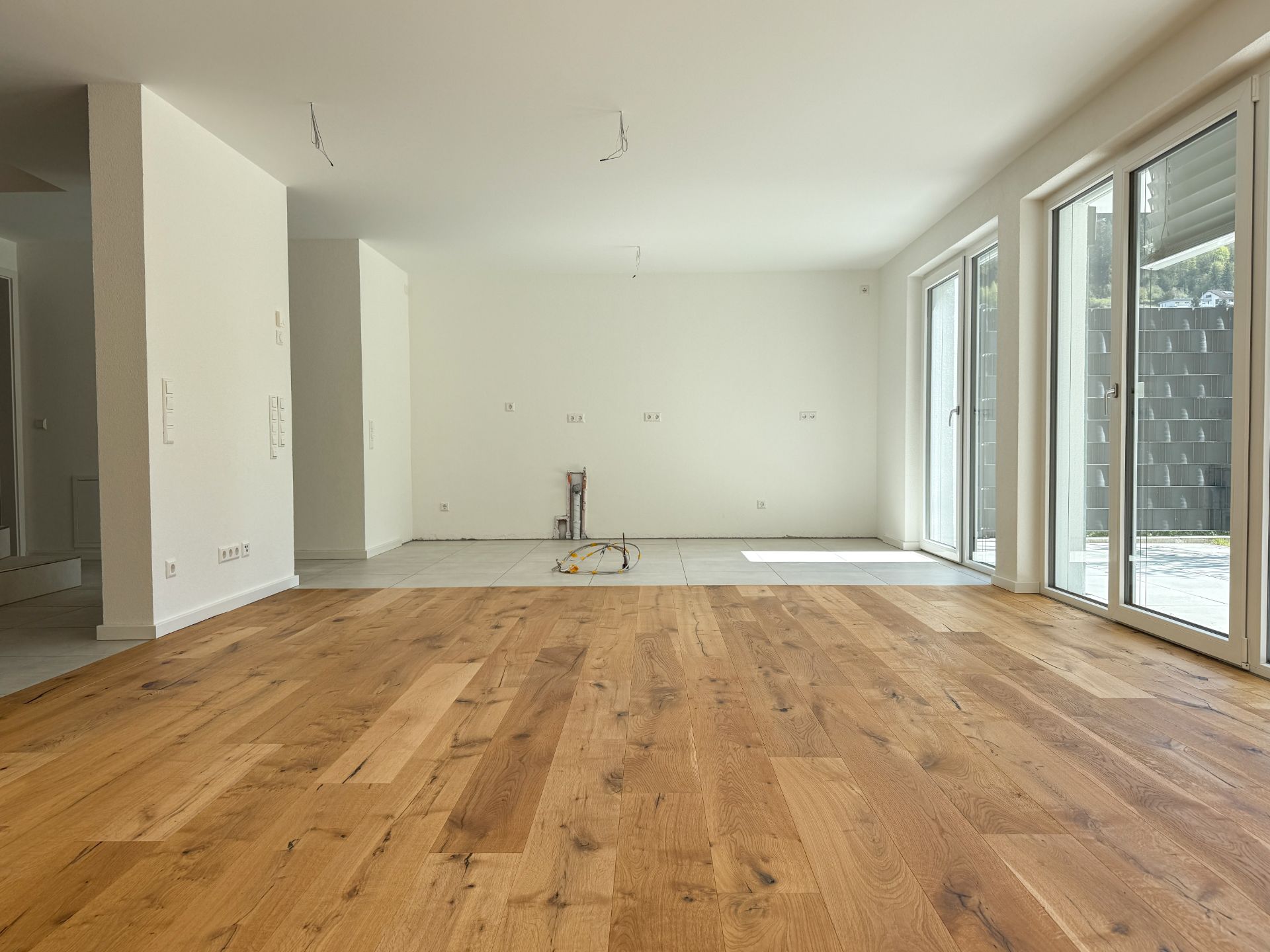 Leeres Wohnzimmer mit Parkettboden, großen Fenstern und Zugang zur Terrasse.
